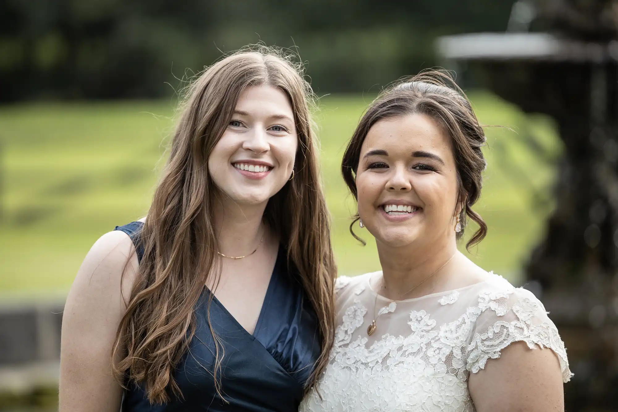 Two women smiling, one in a navy dress and the other in a white lace dress, standing outdoors with greenery and a blurred background.