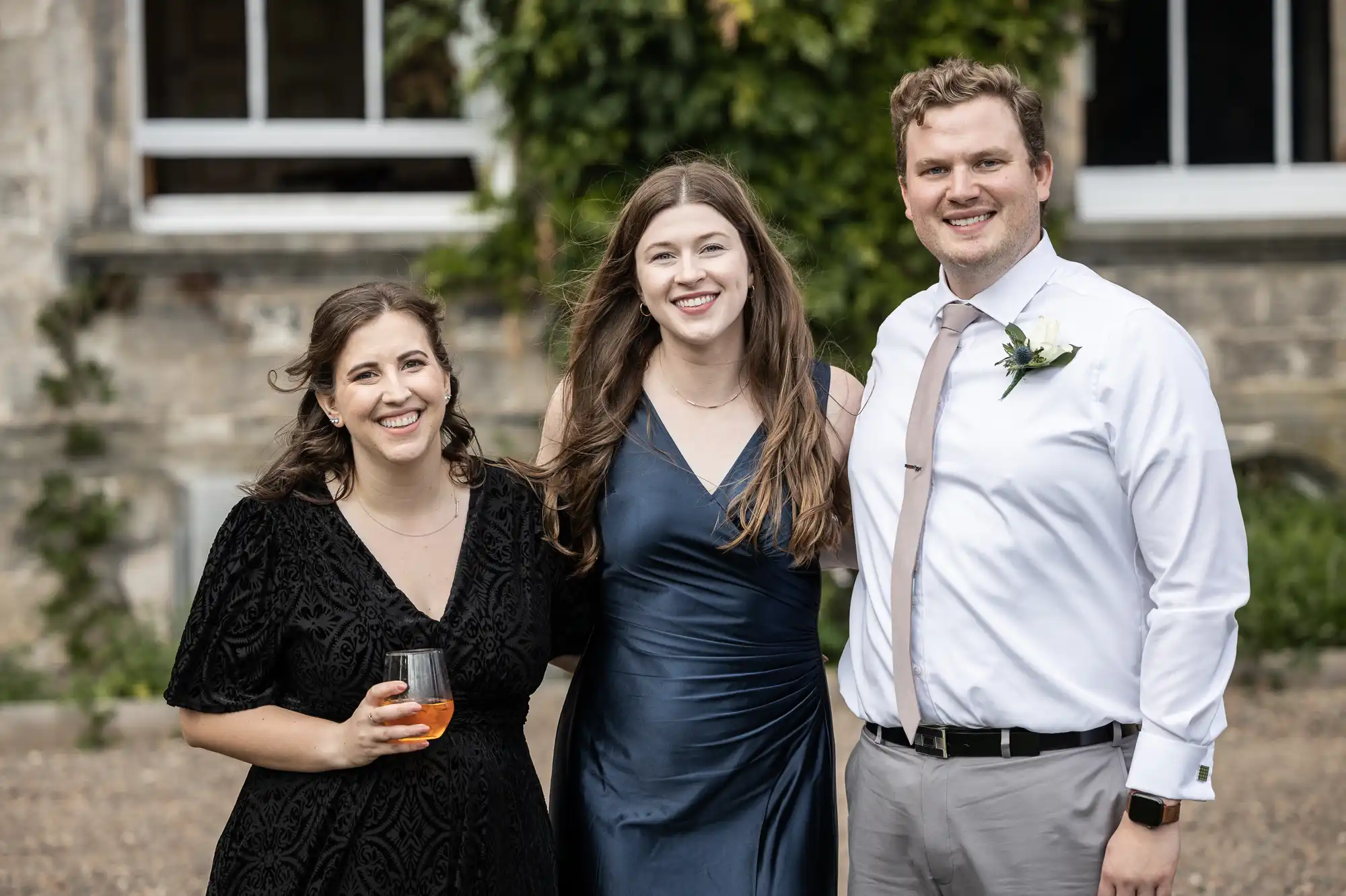 Three people stand together outdoors, smiling at the camera. The person on the left holds a drink, and the person on the right wears a white shirt with a tie and boutonniere.