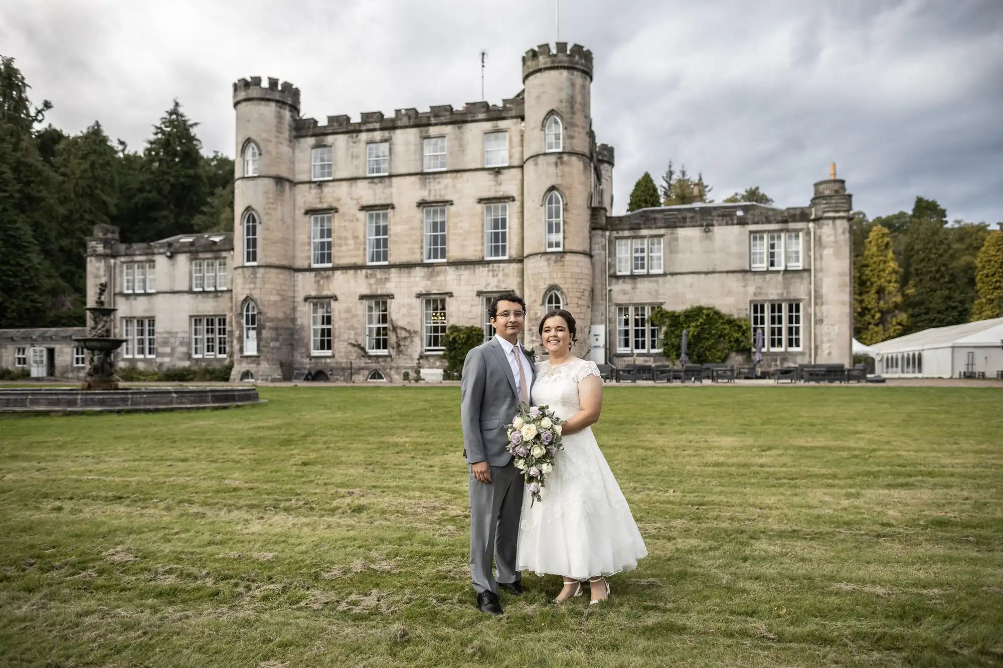 A couple in wedding attire stands on a lawn in front of a large, castle-like building. The groom wears a grey suit while the bride wears a white dress and holds a bouquet.