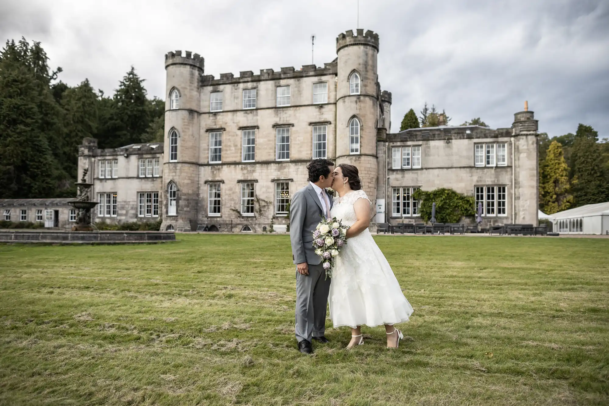 A couple dressed in wedding attire stand close together in front of a large, old castle-like building on a cloudy day. The bride holds a bouquet of flowers.