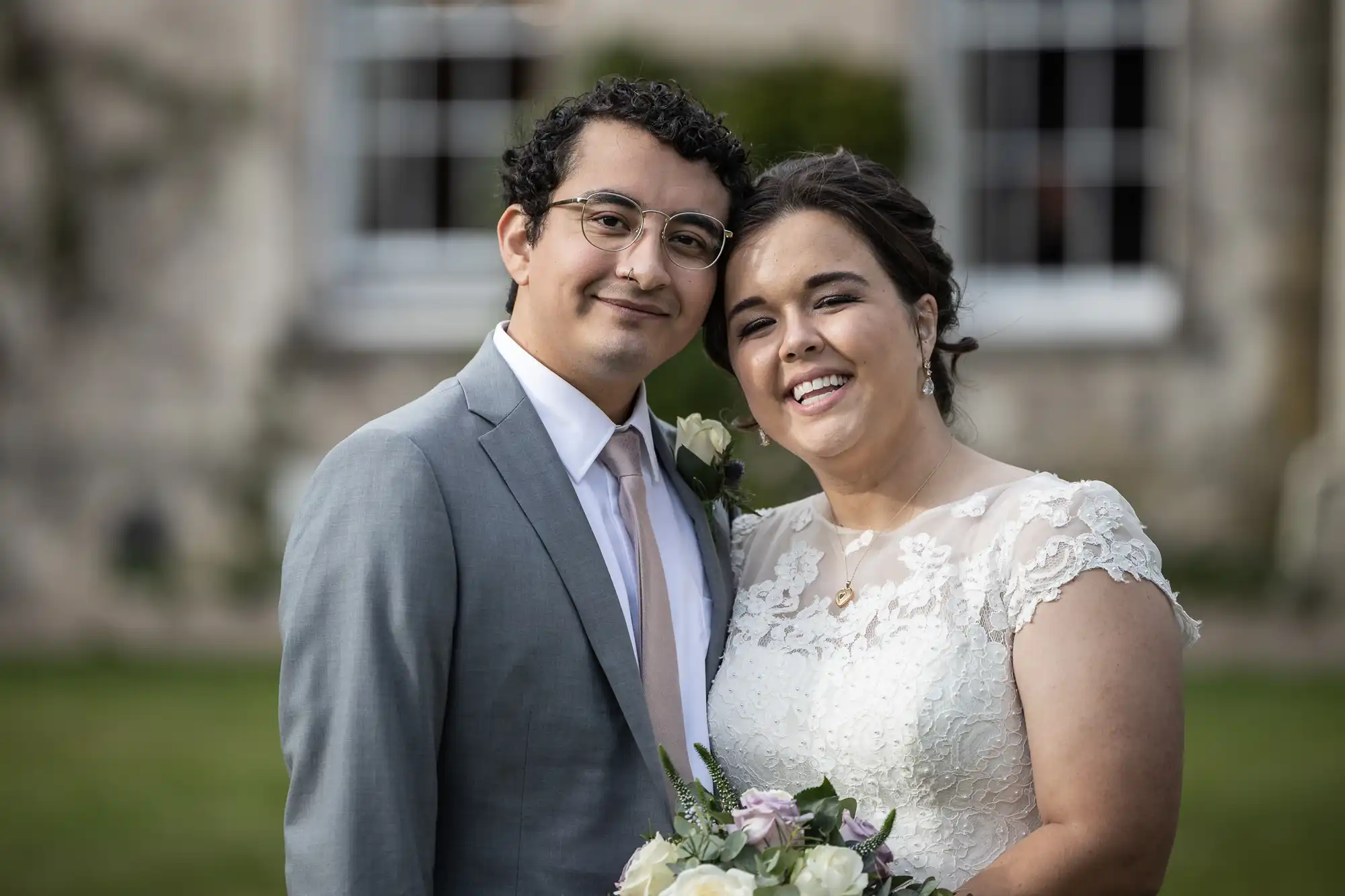 A couple wearing wedding attire smile while posing outdoors, with the woman holding a bouquet of flowers.