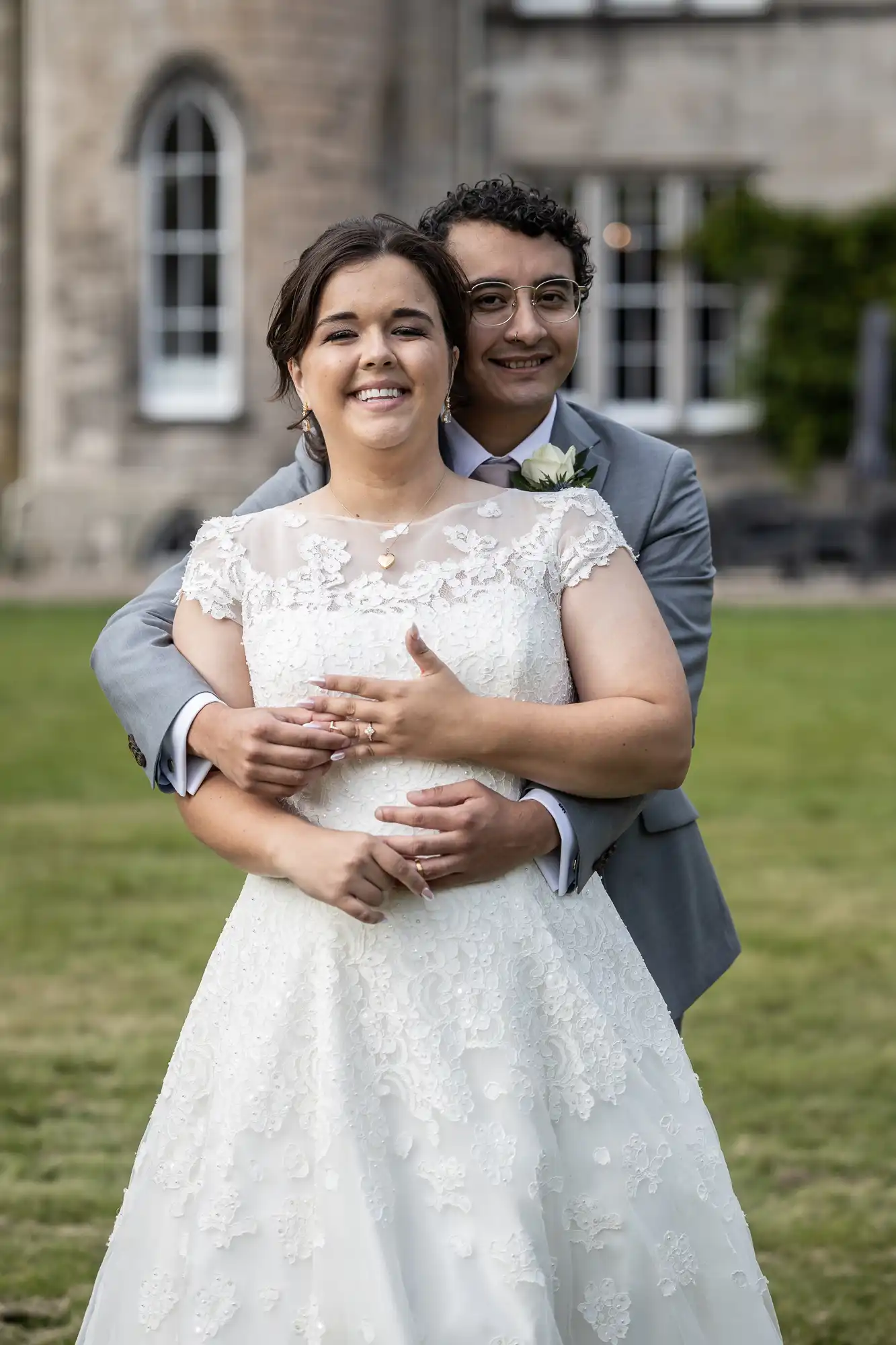 A couple in wedding attire smiles as they embrace outdoors, with a stone building and lawn in the background.