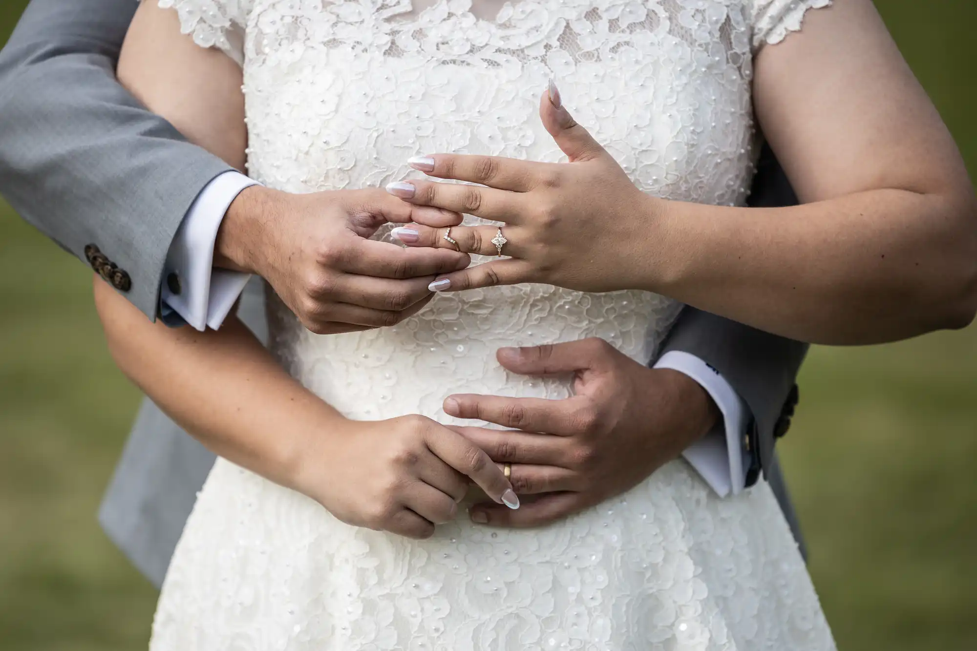 A couple embraces from behind, highlighting the bride's lace wedding dress and her engagement ring.