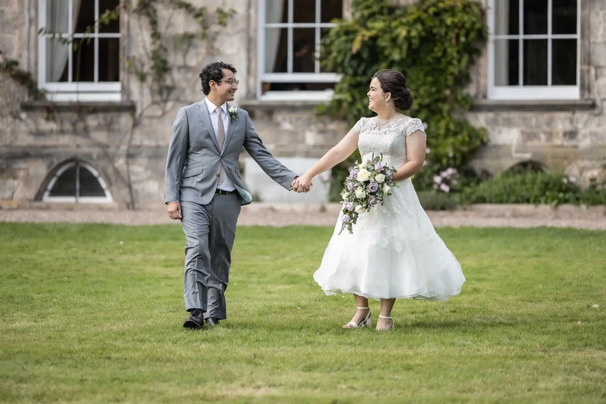 A bride and groom hold hands and smile at each other while walking on grass in front of a stone building with large windows and greenery. The bride holds a bouquet of flowers.