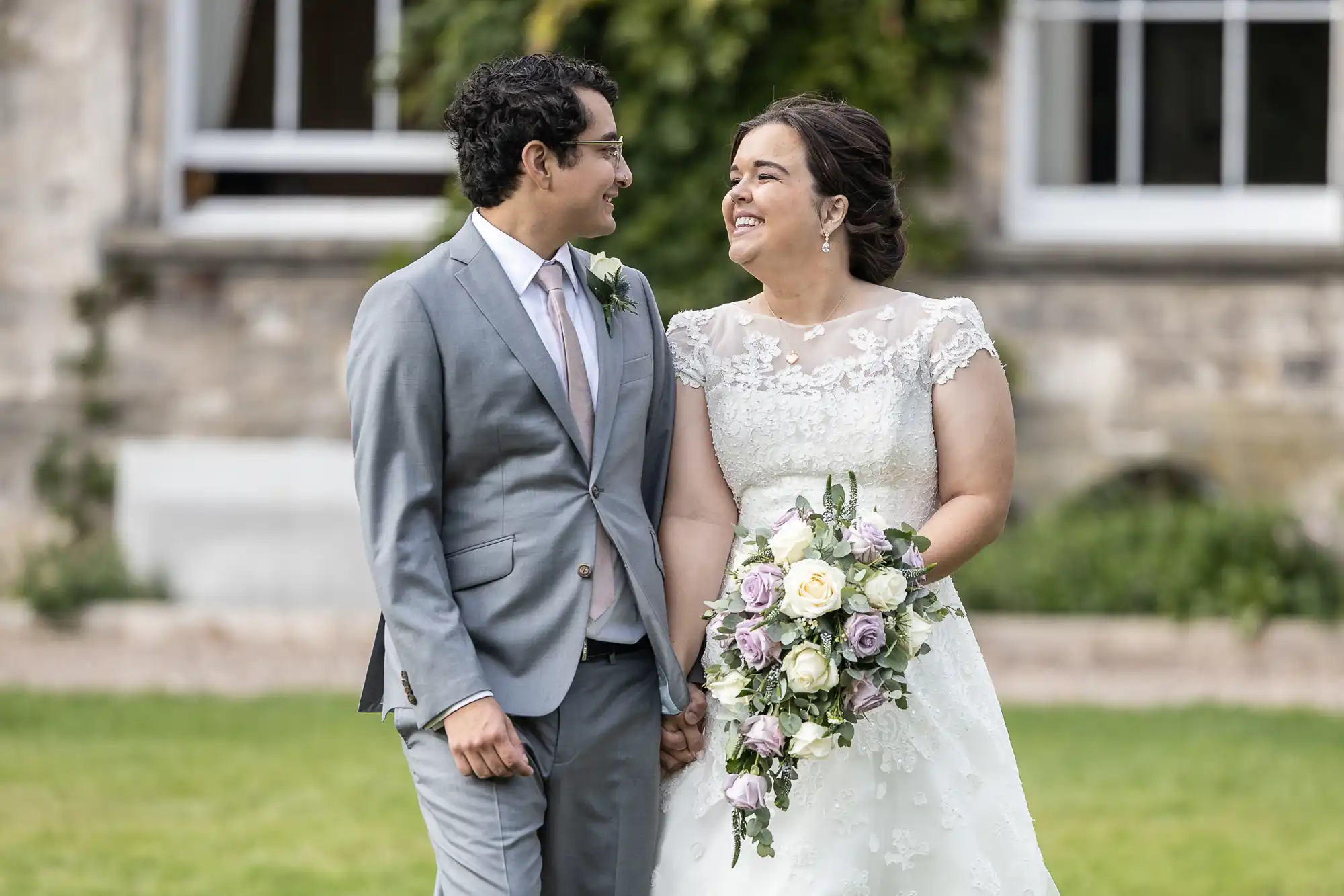 A couple is standing outdoors, smiling at each other. The woman is wearing a white wedding dress and holding a bouquet, while the man is in a light grey suit.