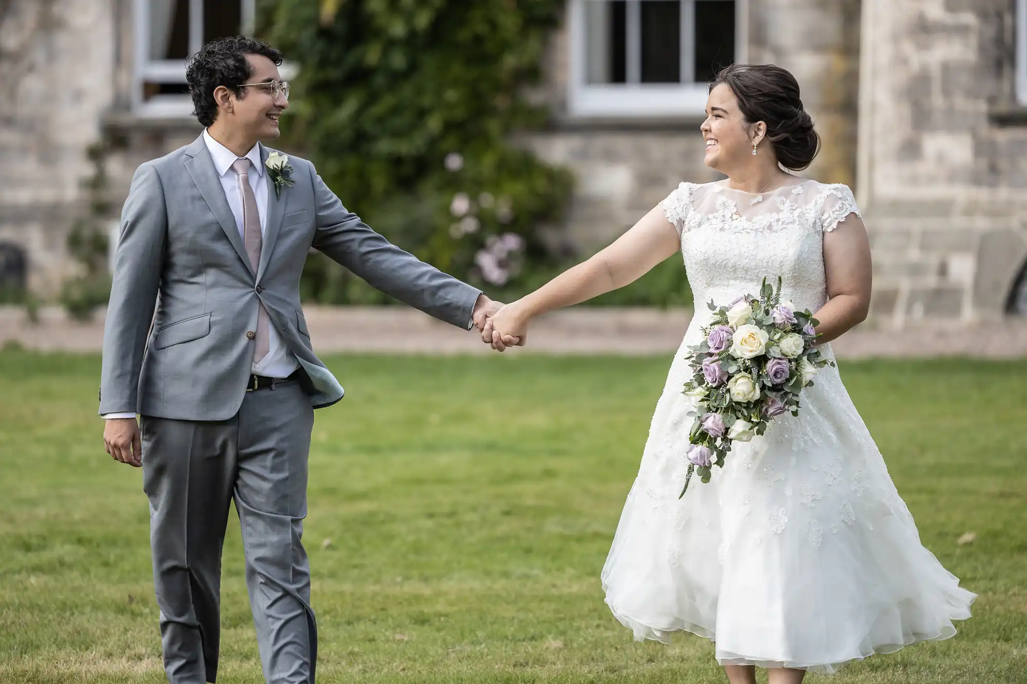 A bride and groom hold hands while walking on grass in front of a building. The bride wears a white dress and holds a bouquet, and the groom wears a gray suit with a pink tie.