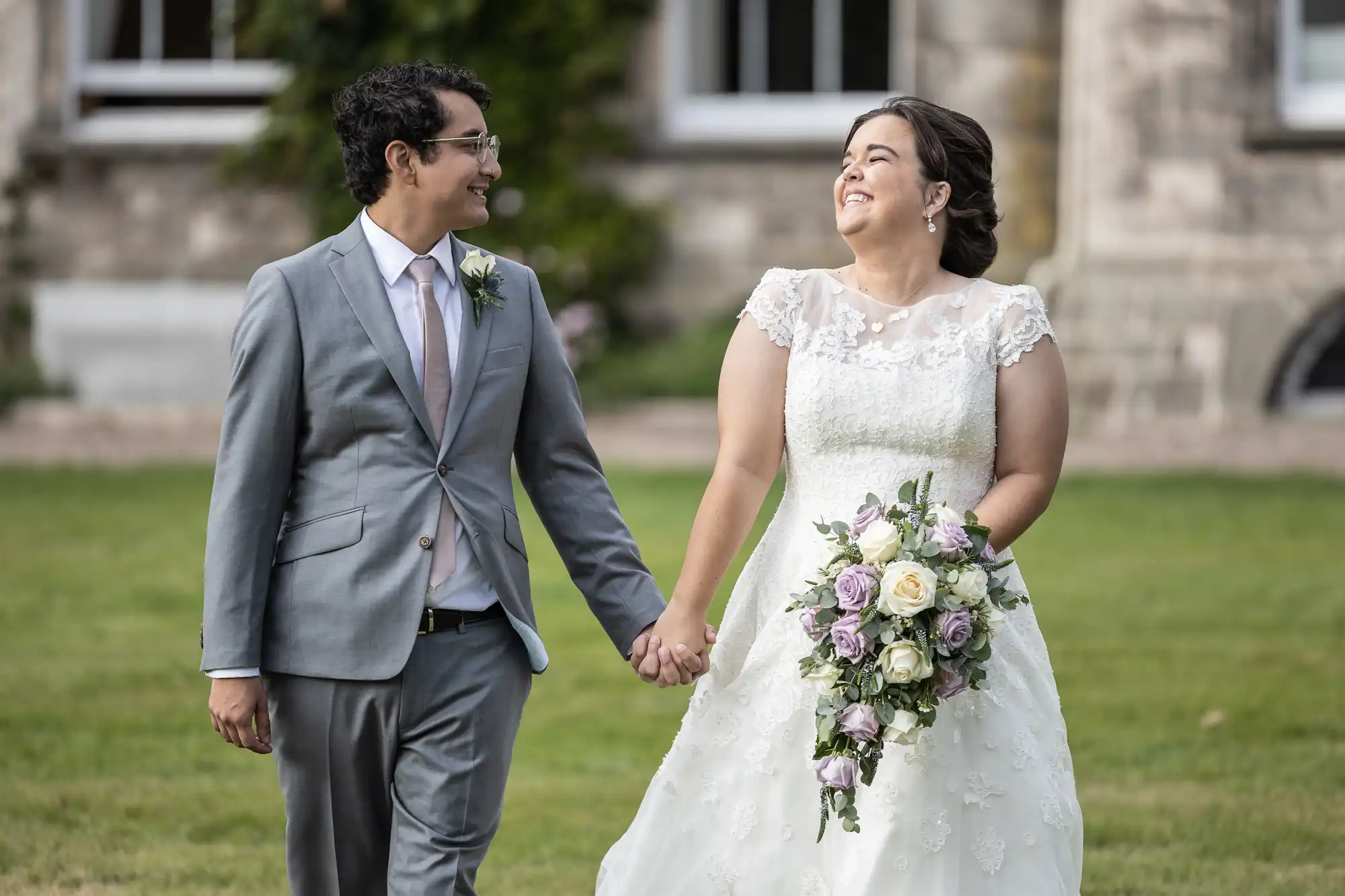 A couple holding hands and smiling at each other, dressed in wedding attire, with a bouquet of flowers, walking on a lawn in front of a stone building.