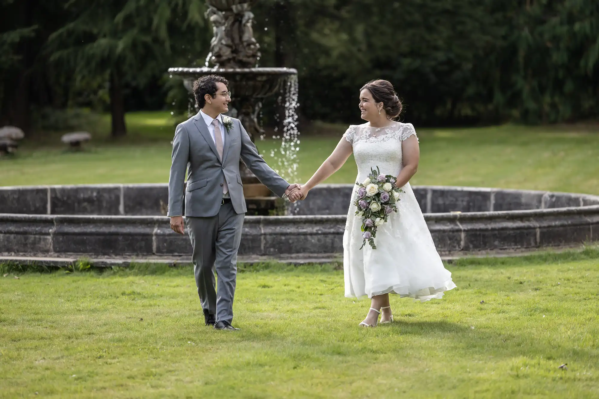 A bride and groom walk hand in hand on a grassy lawn in front of a stone fountain. The groom wears a gray suit and tie, while the bride is in a white dress holding a bouquet of flowers.