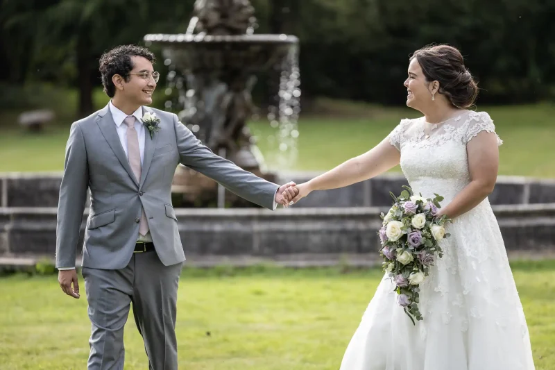 Melville Castle wedding photos: A couple in wedding attire holds hands and smiles at each other. The groom is in a grey suit and the bride in a white dress. They stand in front of a fountain in a green outdoor setting.