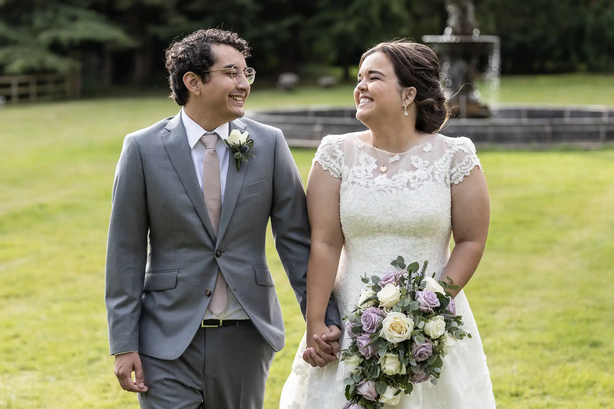 A bride and groom smile at each other while walking hand in hand outdoors. The bride holds a bouquet of white and purple flowers.