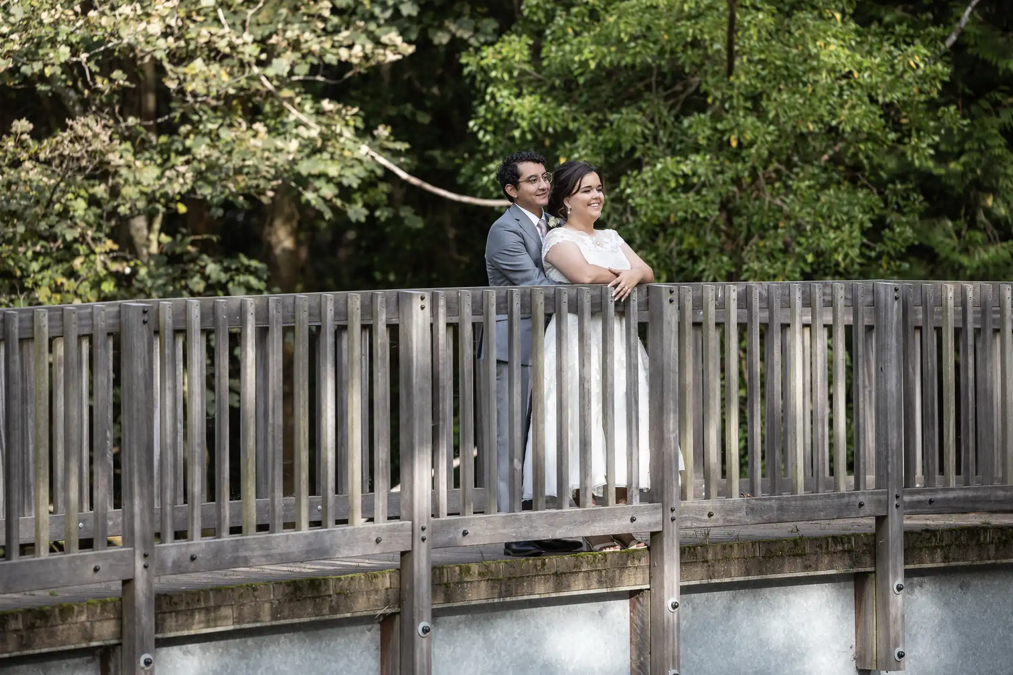 A couple in wedding attire stands closely together on a wooden bridge, surrounded by trees and greenery.