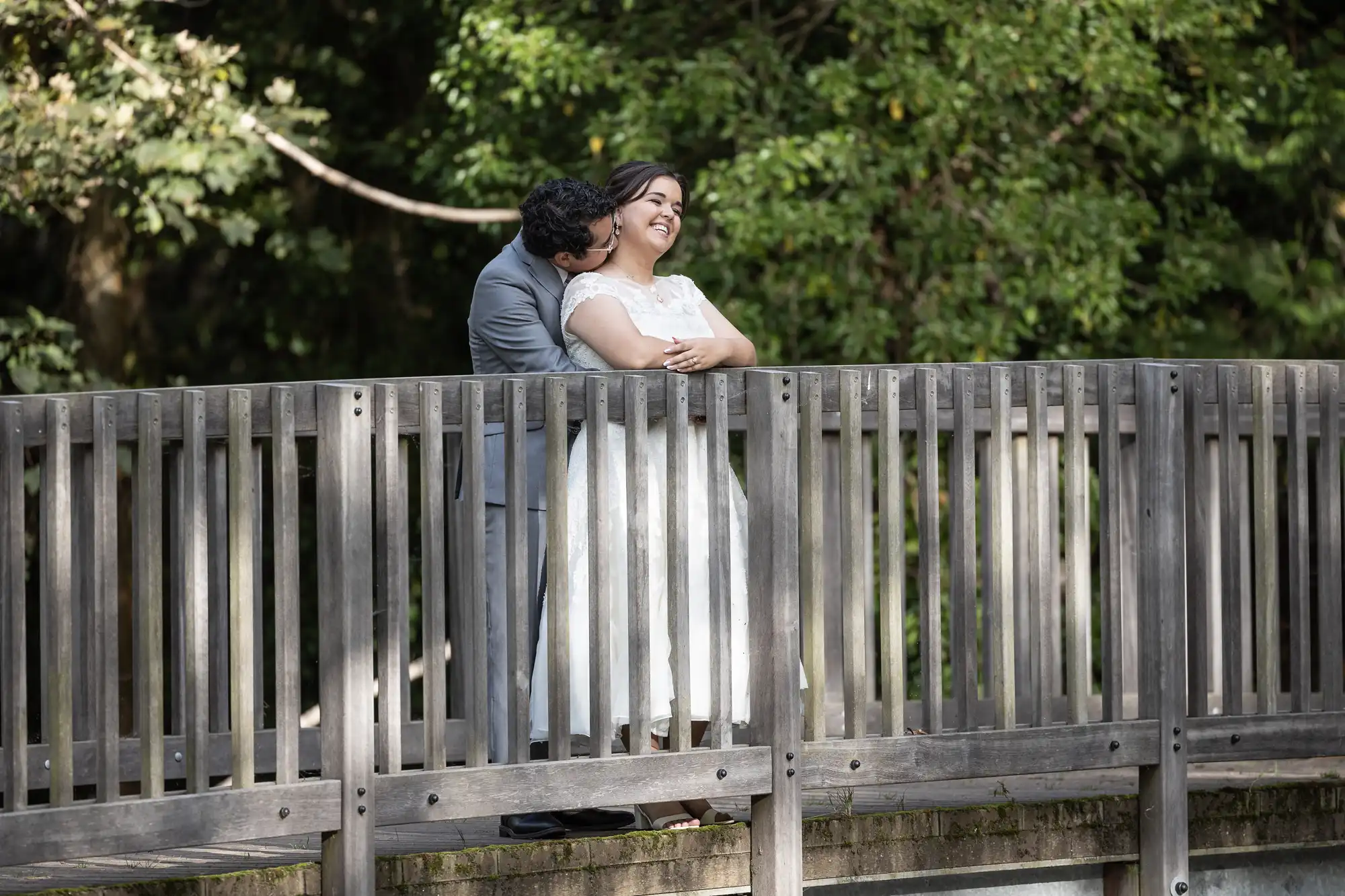 A couple stands on a wooden bridge with the man in a gray suit hugging the woman in a white dress from behind. They are outdoors with green trees in the background.