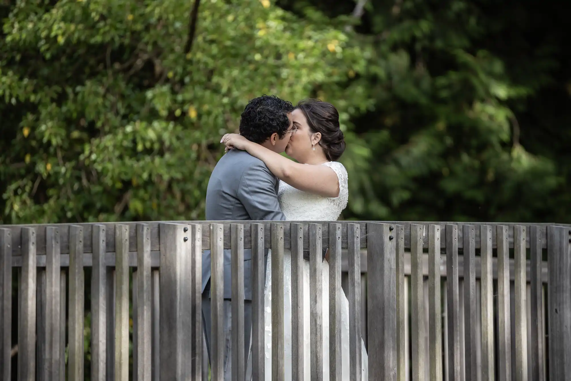 A couple embraces and kisses while standing on a wooden bridge in an outdoor, wooded area.