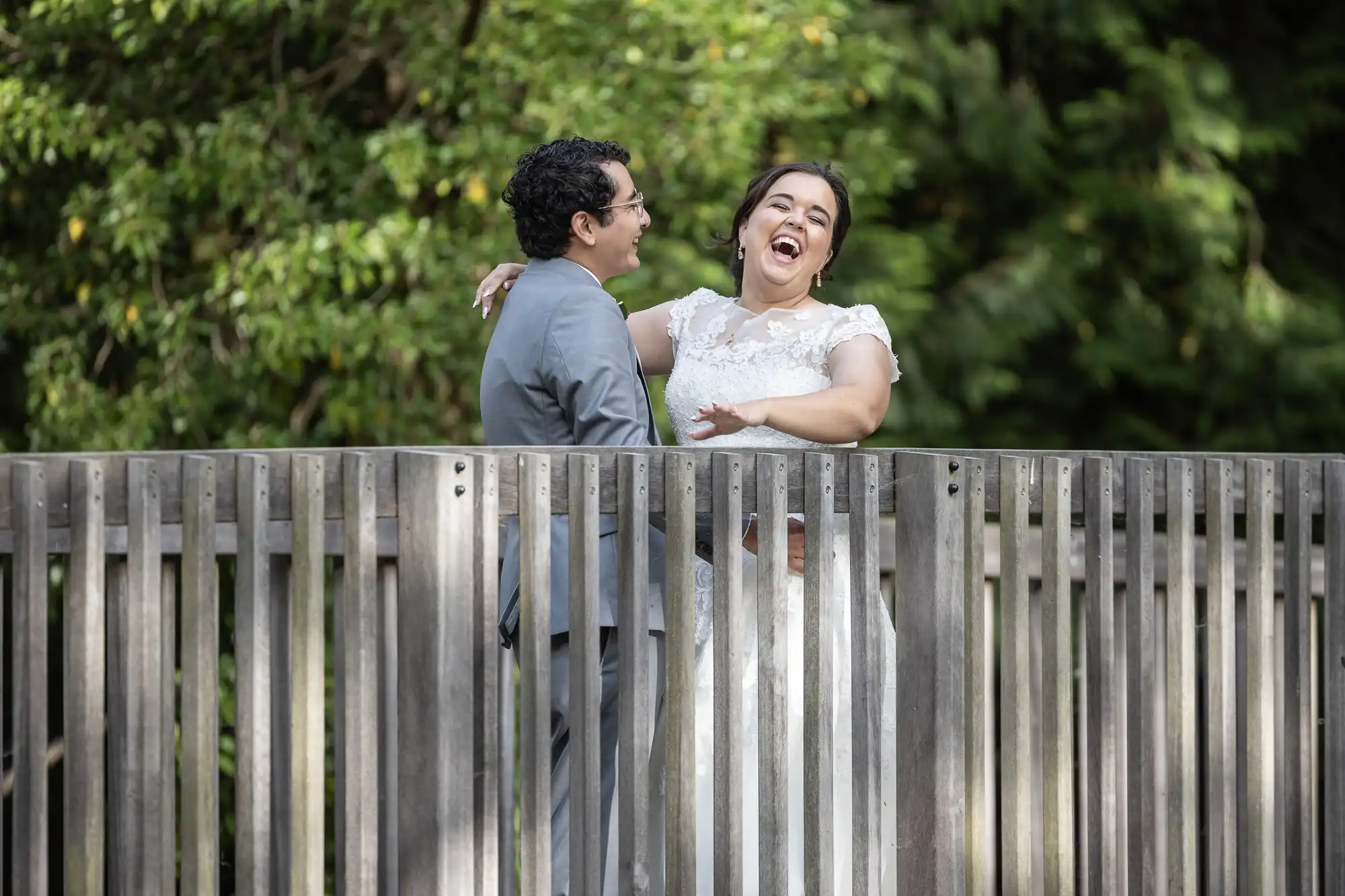 A couple stands on a wooden bridge, smiling and laughing, with greenery in the background.