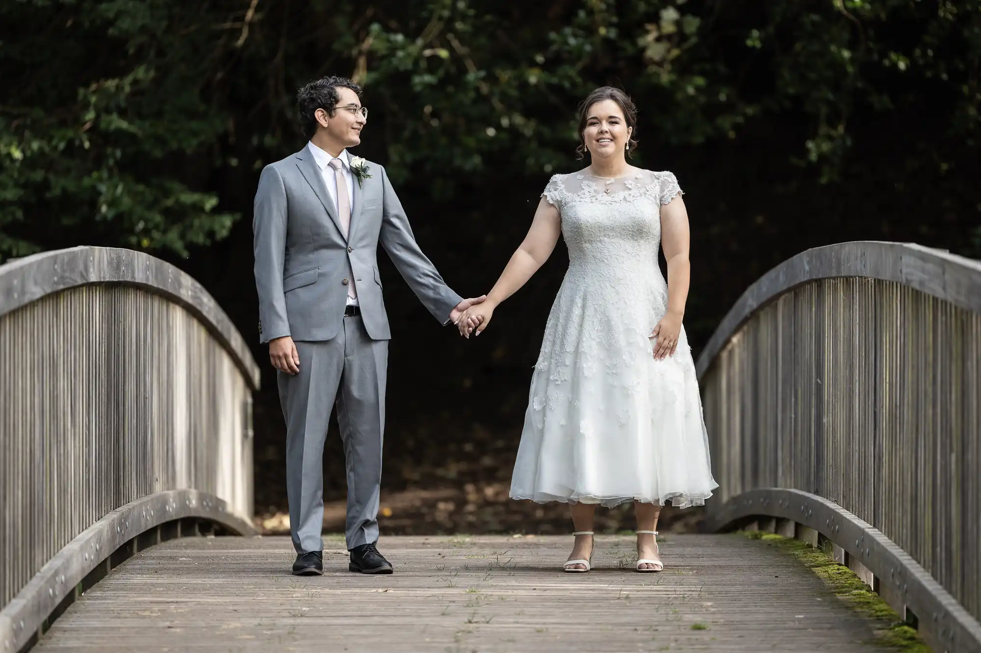 A couple in formal attire, with the man in a gray suit and the woman in a white dress, hold hands while standing on a wooden bridge. Trees are visible in the background.