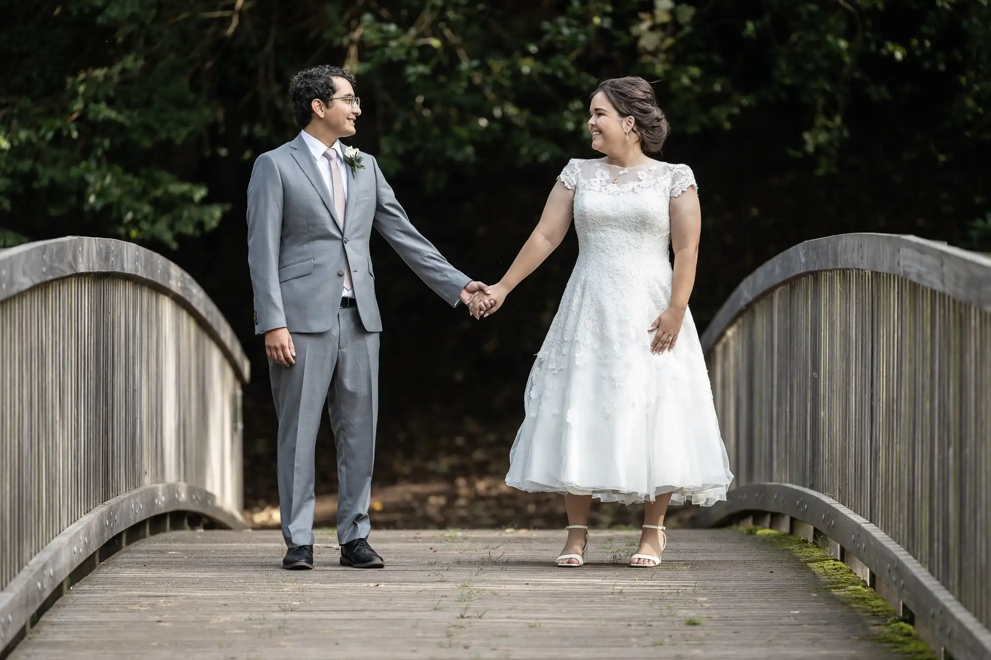 A couple in wedding attire holds hands while standing on a wooden bridge. The groom is in a gray suit, and the bride wears a white dress. They are facing each other with trees in the background.