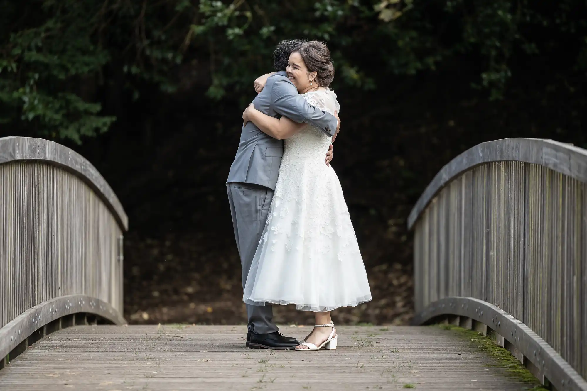 A couple embracing on a wooden bridge, with the bride in a white dress and the groom in a gray suit. Trees are visible in the background.