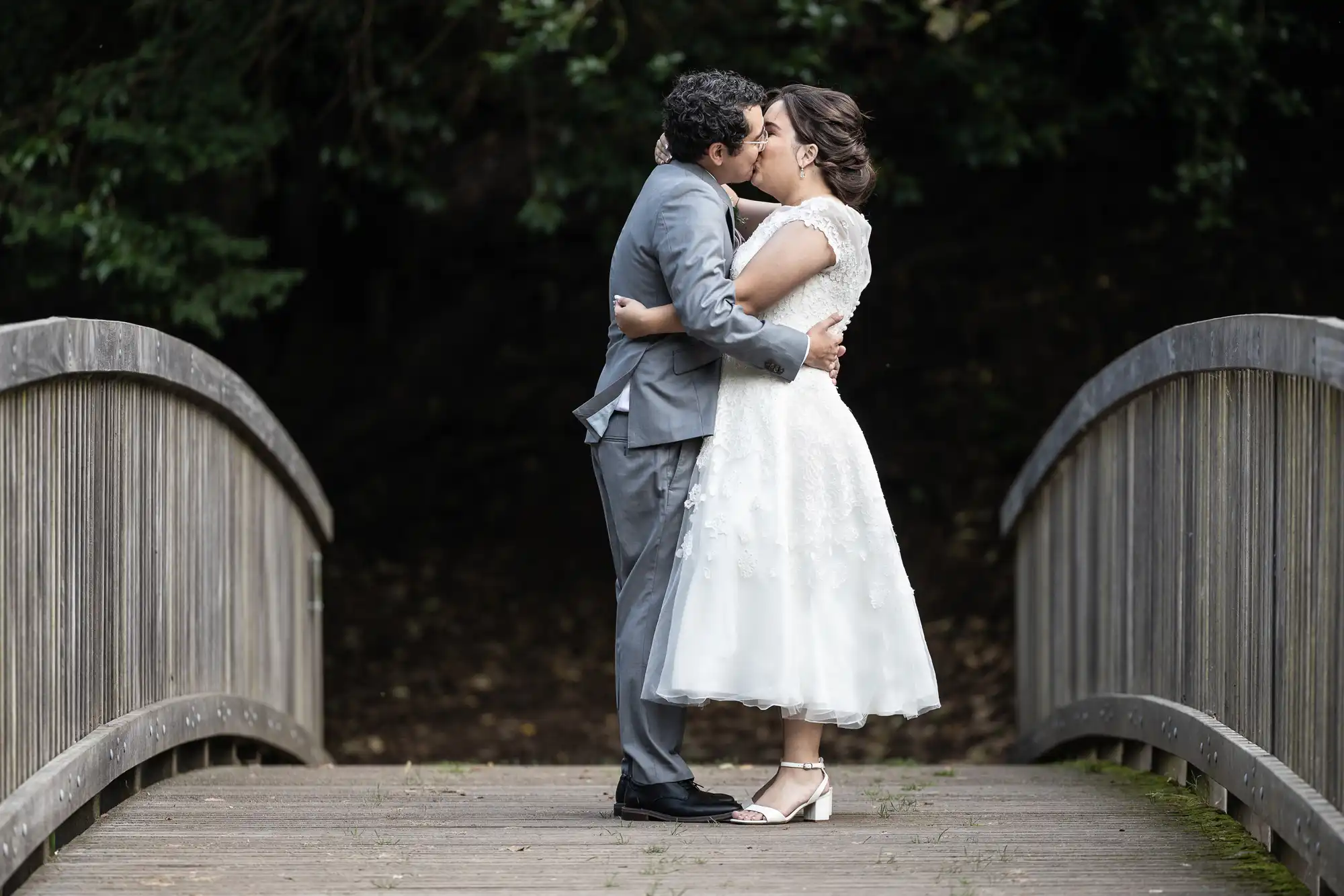 A couple in wedding attire shares a kiss while standing on a wooden bridge surrounded by greenery.