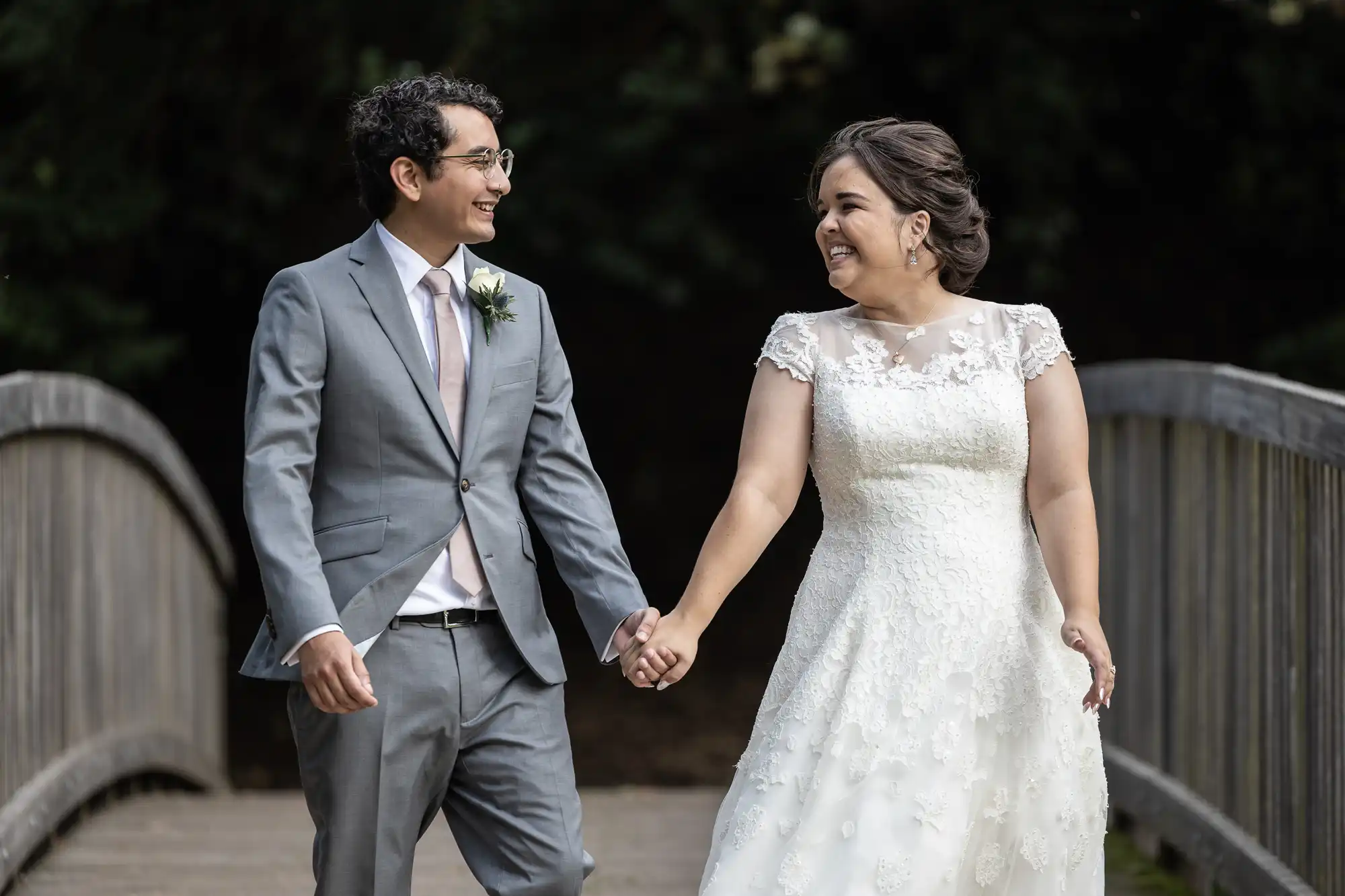 A couple holding hands and smiling at each other on a wooden bridge, the man in a gray suit and the woman in a white wedding dress.