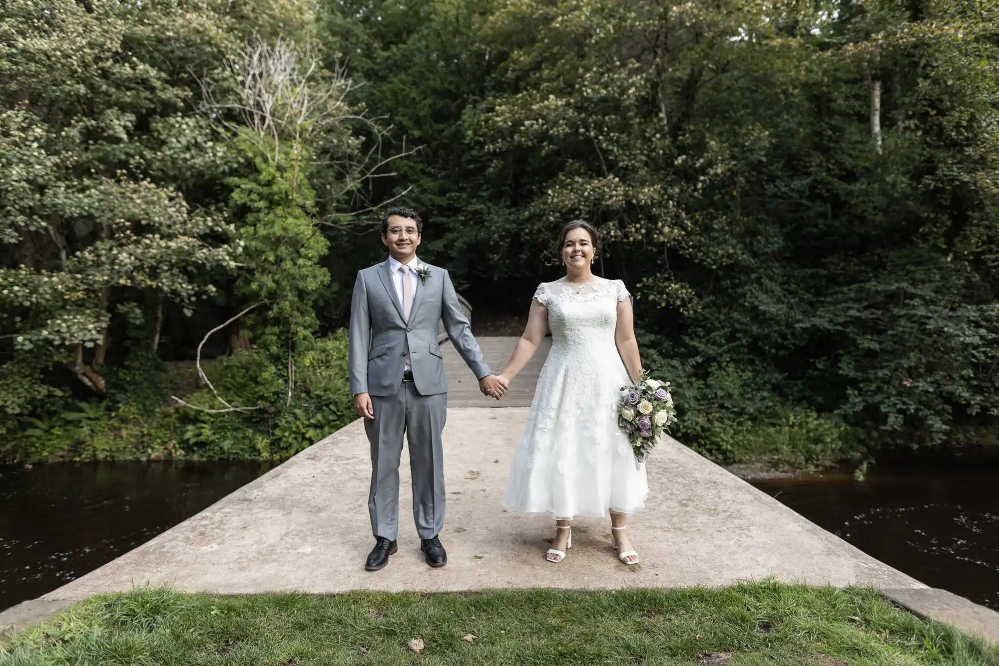 A couple holding hands, standing on a concrete path near a river with dense greenery in the background. The groom is in a gray suit while the bride is in a white dress holding a floral bouquet.