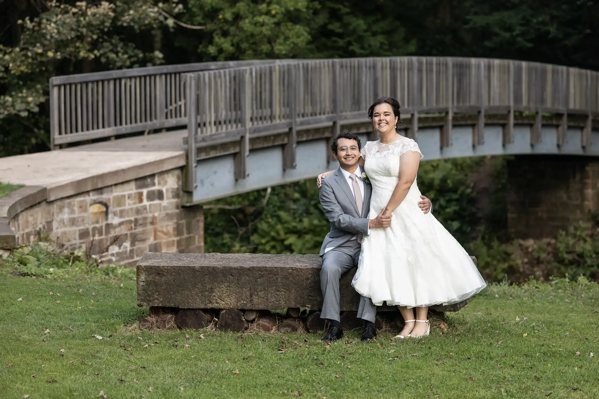 A couple, with the woman in a white dress and the man in a gray suit, poses sitting on a bench in front of a wooden bridge in a park setting.