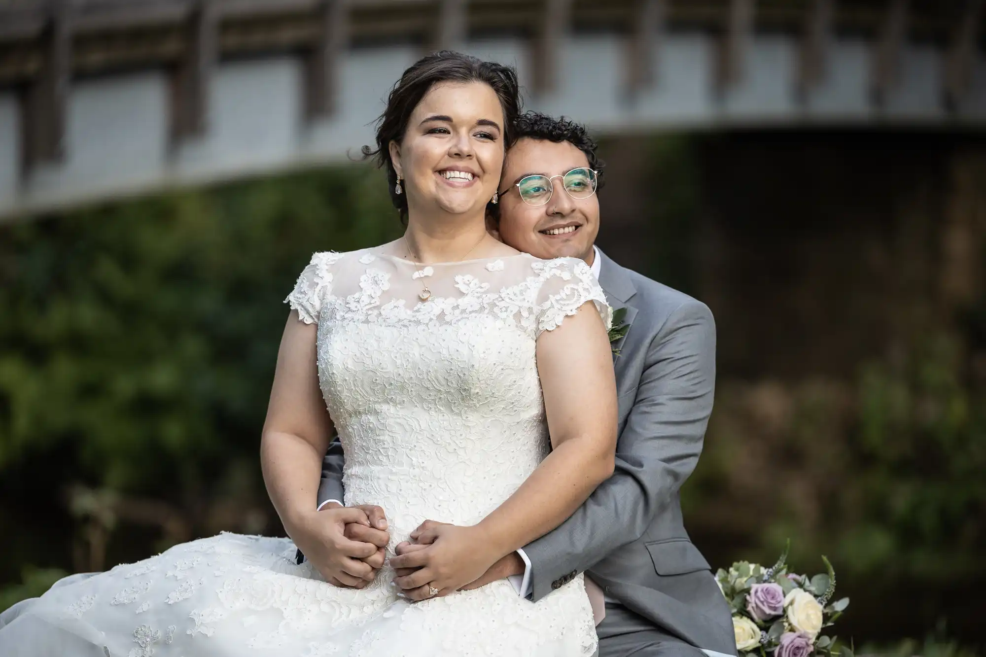 Bride in a white lace wedding dress and groom in a gray suit with glasses sit closely together, smiling. A bouquet of flowers is placed nearby. They are outdoors in front of a blurred bridge and foliage.