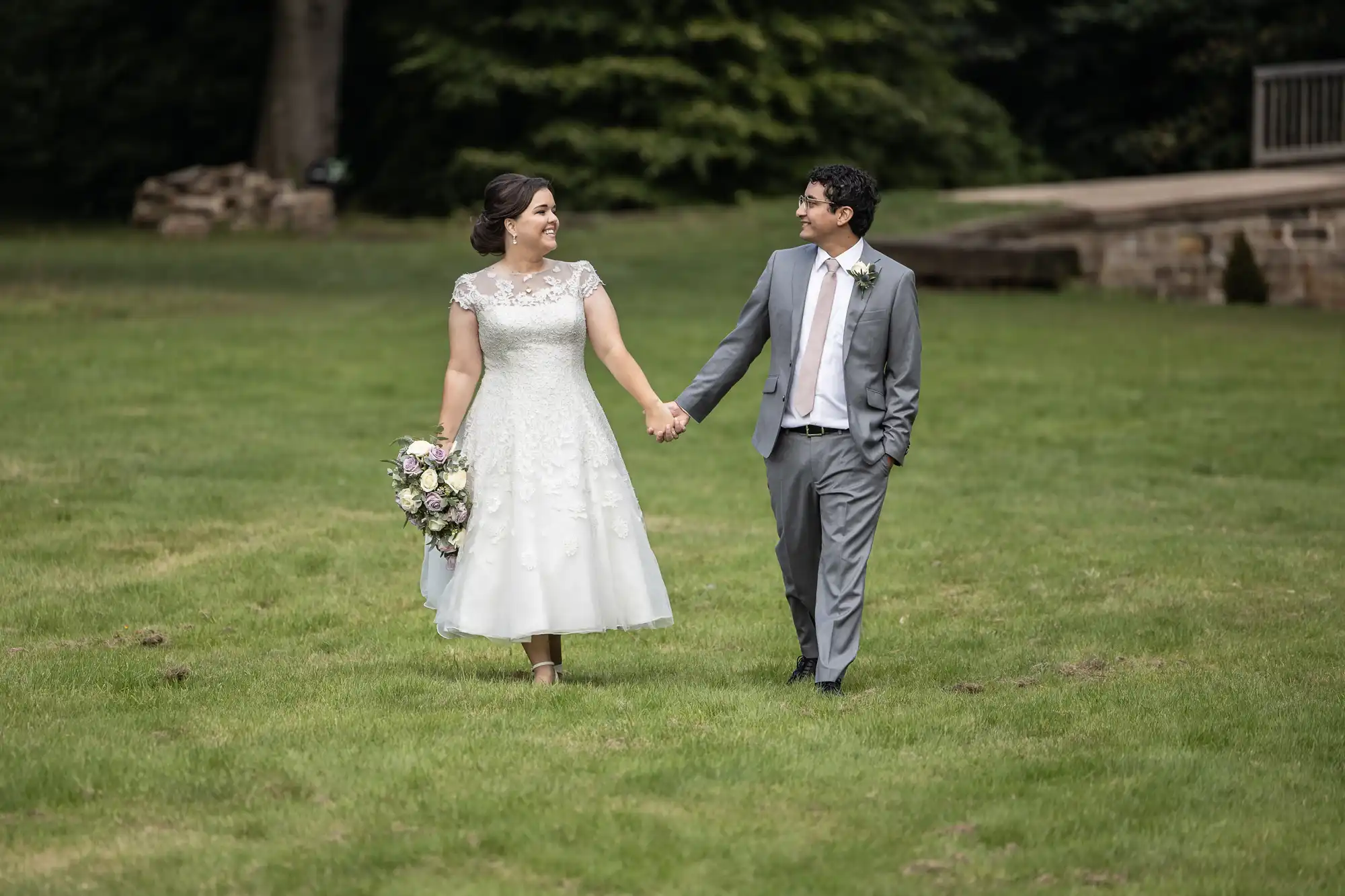 A bride in a white dress and a groom in a gray suit walk hand-in-hand on a grassy lawn, both smiling. The bride is holding a bouquet of flowers. Trees and a wooden structure are in the background.