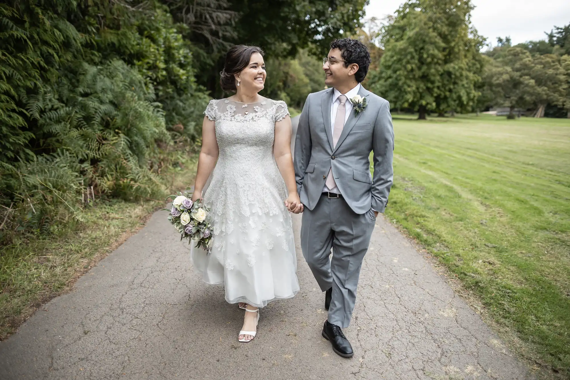 A bride in a white dress and a groom in a gray suit walk hand in hand on a path, smiling and looking at each other, with greenery in the background.