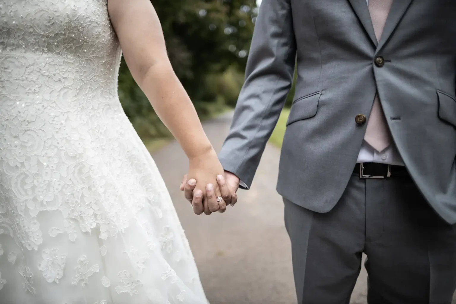A bride in a white lace wedding dress and a groom in a gray suit hold hands while standing on a path outdoors.