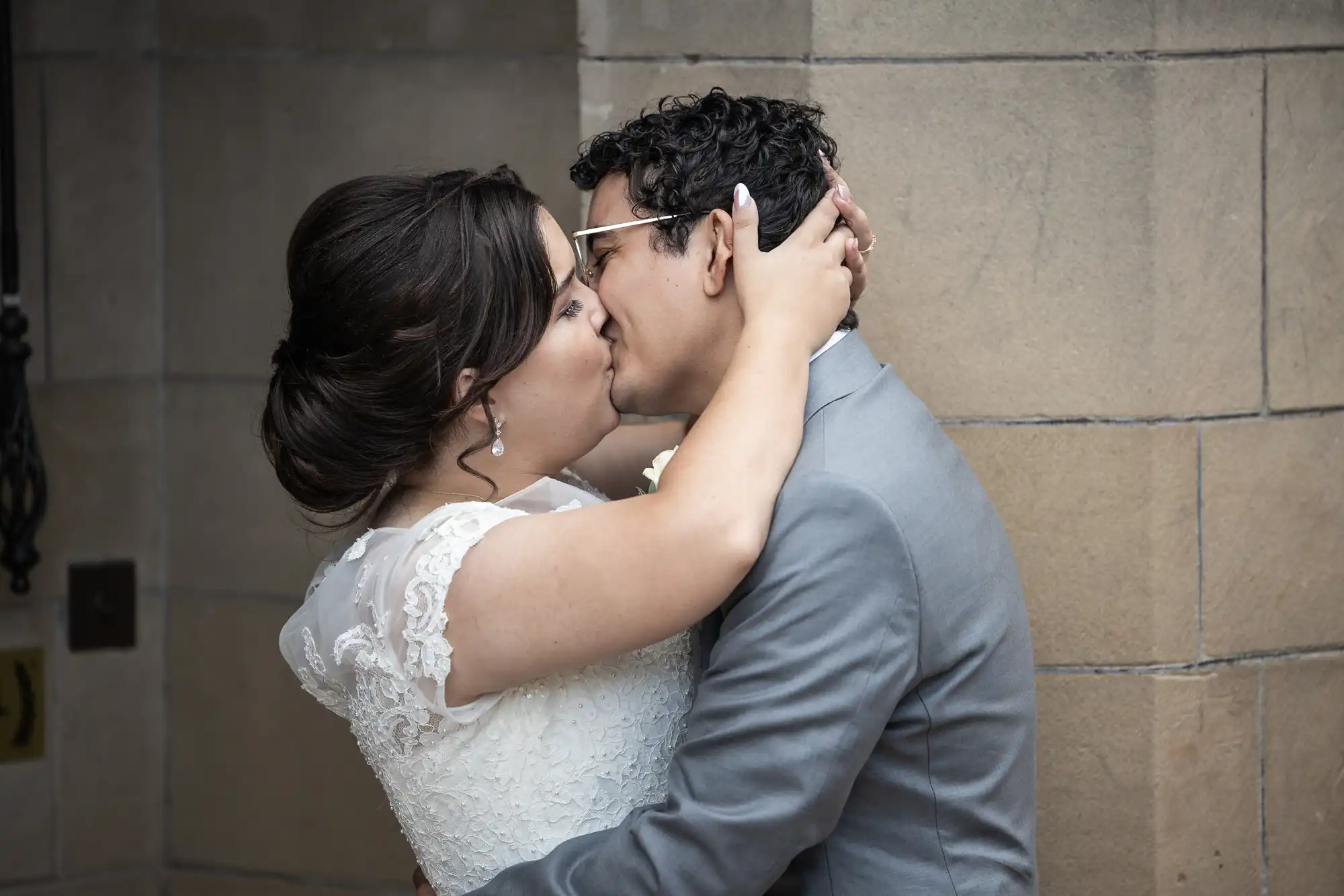 A bride and groom embrace and kiss while standing against a stone wall. The bride wears a white lace dress, and the groom is in a light gray suit.