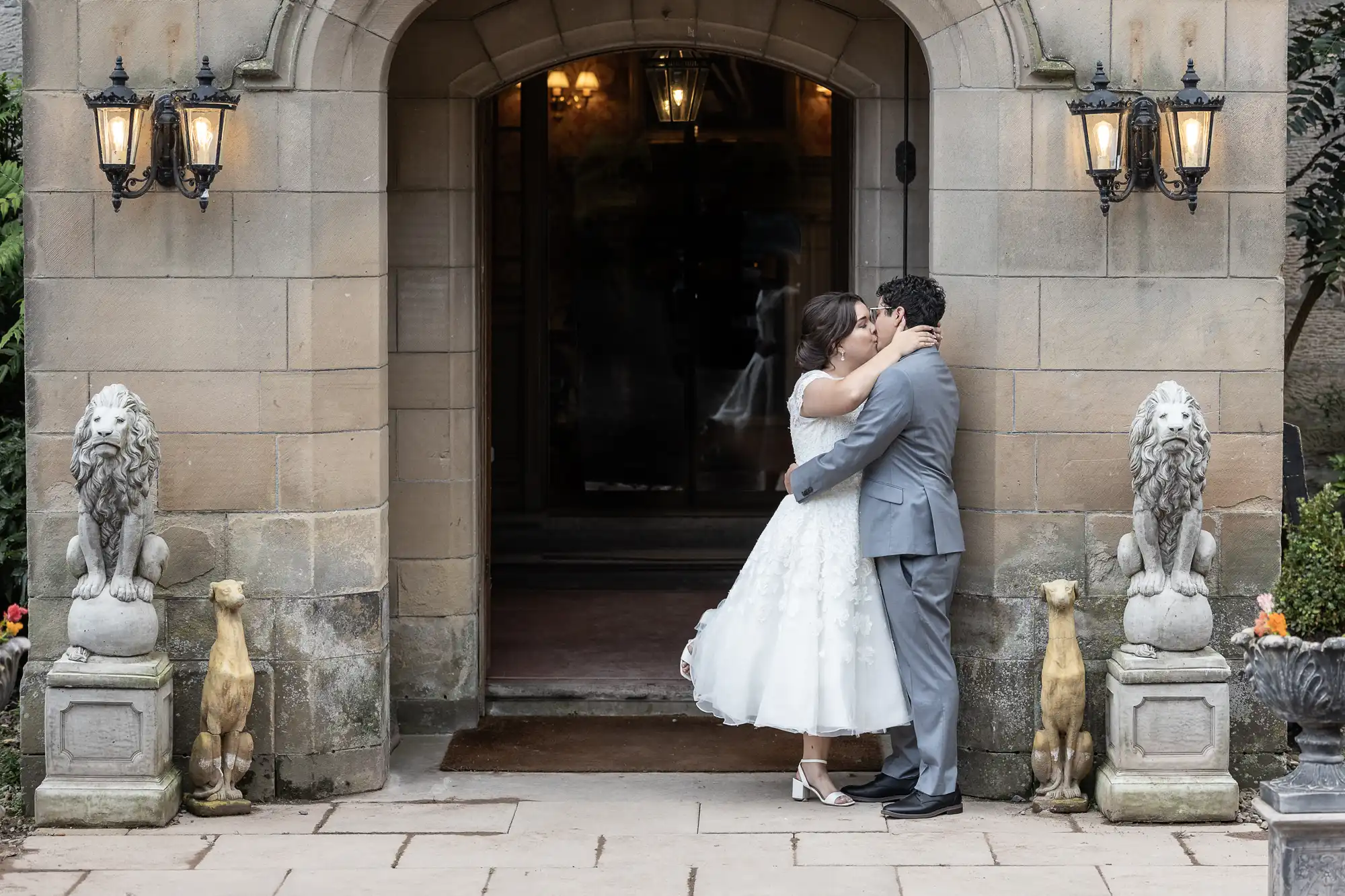 A bride and groom share a kiss in front of an arched stone doorway flanked by stone lion statues. The groom wears a gray suit, and the bride wears a white dress. Four lanterns are mounted on the doorway.