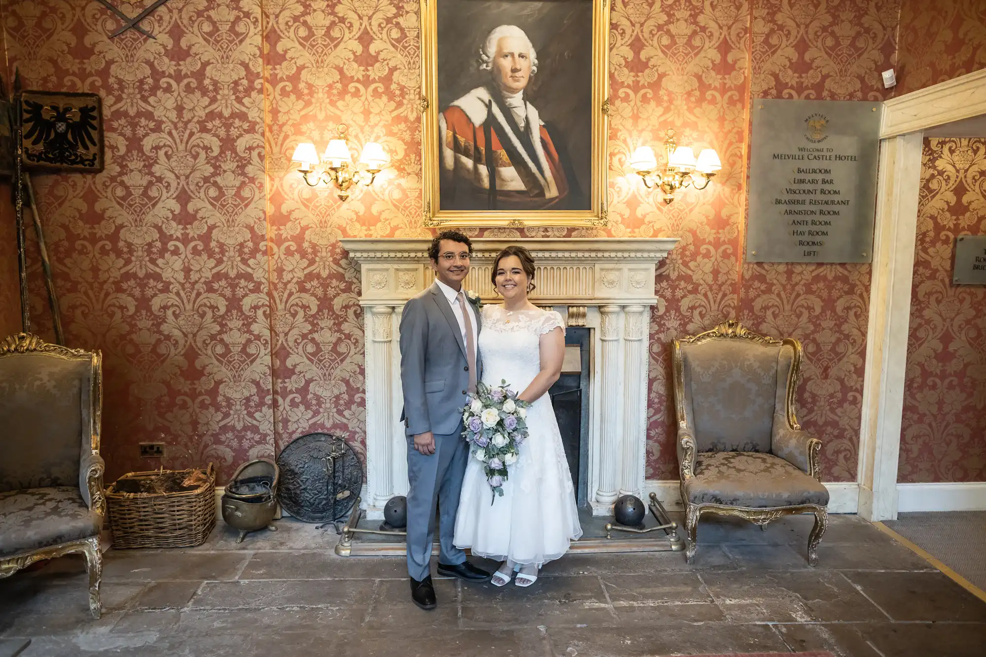 A couple, dressed in formal wedding attire, stand smiling in front of a fireplace. A large portrait and a brass plaque with text are mounted on the textured, red patterned wall behind them.