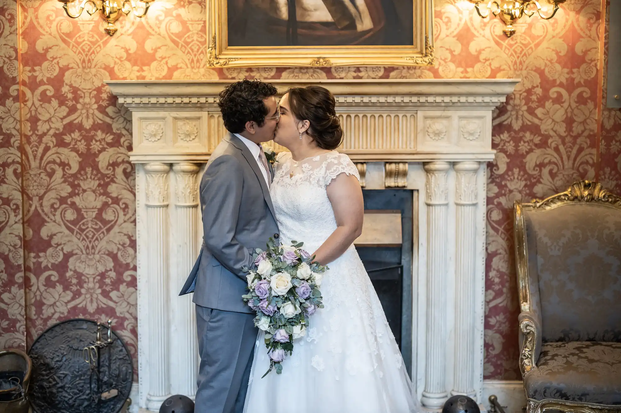 A bride and groom kiss in front of a white fireplace with an ornate gold and red wallpaper behind them. The bride holds a bouquet of purple and white flowers.