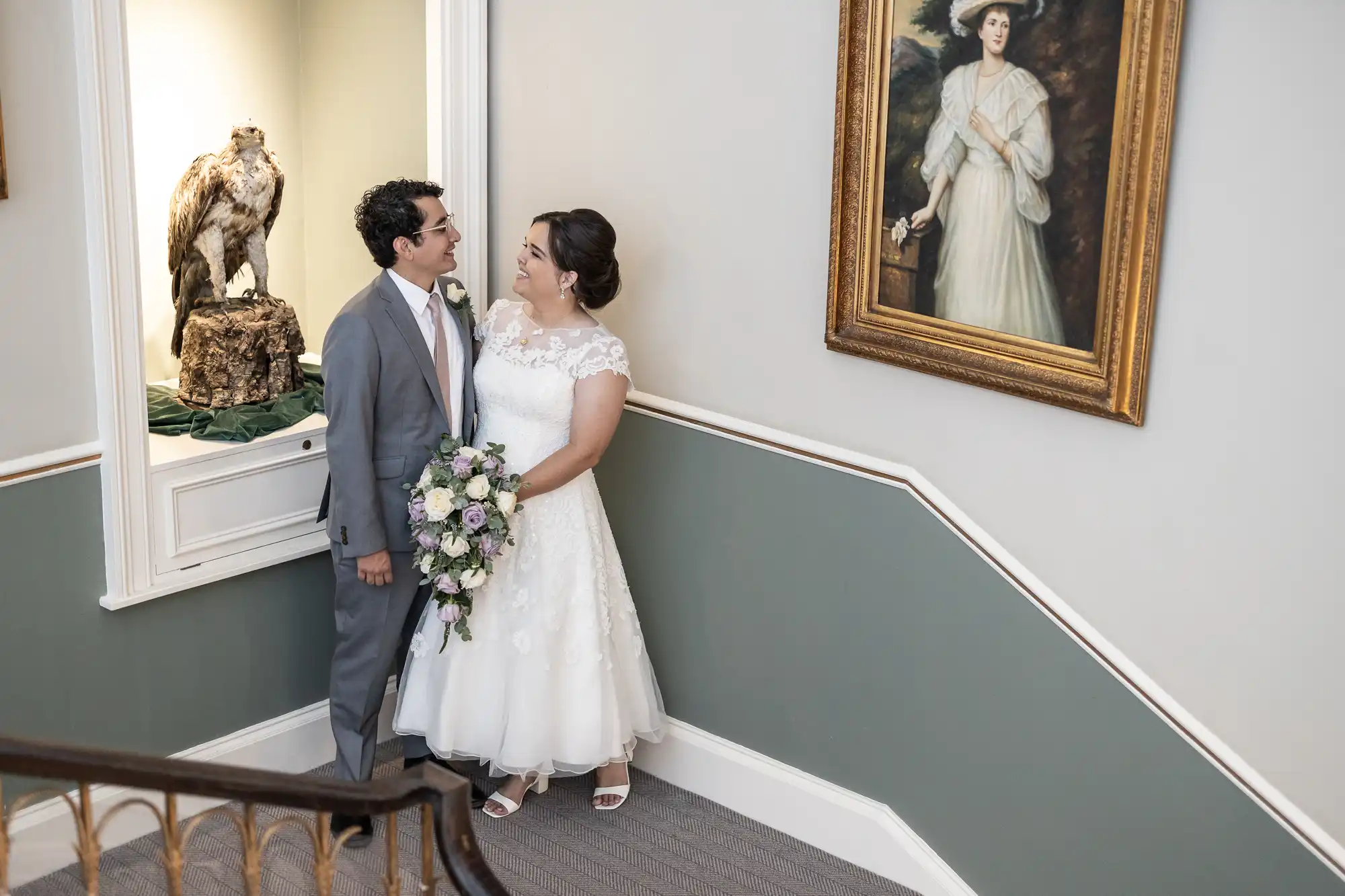 A couple on their wedding day stands at the foot of a staircase beside a bird display and a portrait of a woman. The bride holds a floral bouquet, and both are dressed in wedding attire.