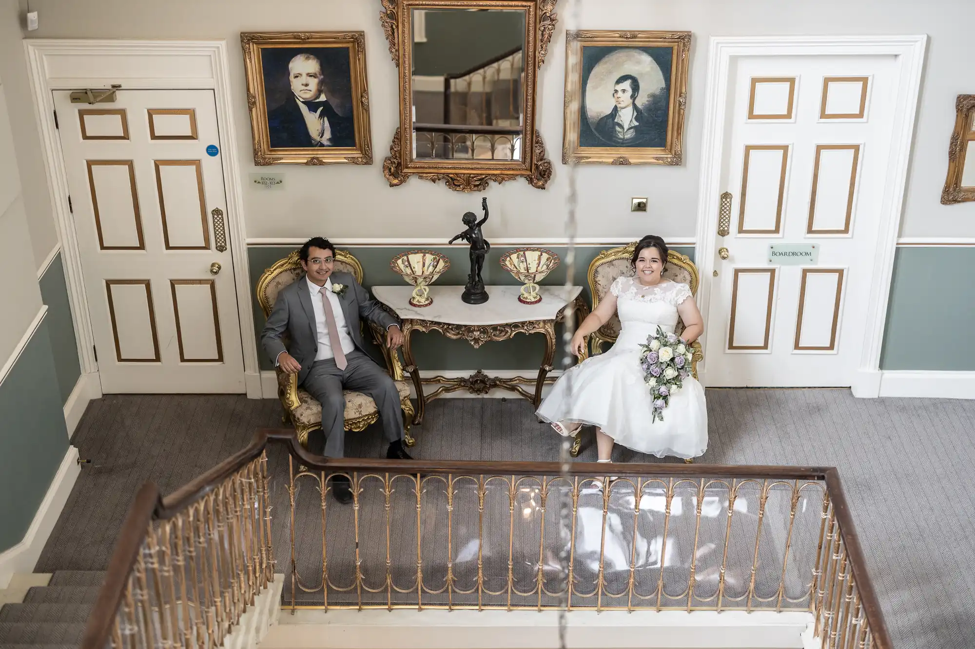 A couple in formal attire sits in ornate chairs in a vintage room with framed portraits and a mirror on the wall. The woman holds a flower bouquet.