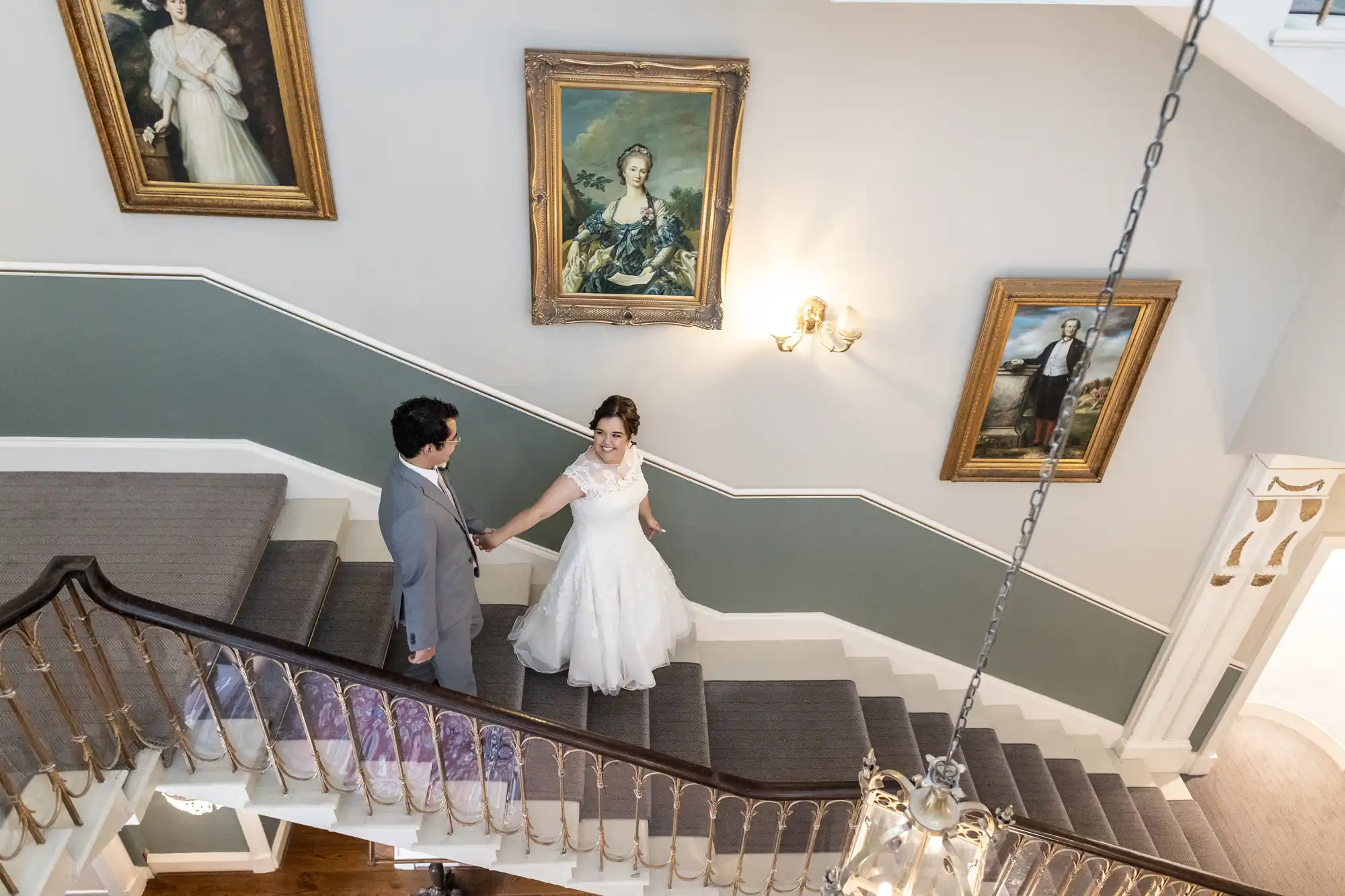 A couple in wedding attire descends a staircase in a gallery adorned with classical paintings. The bride holds the groom's hand as they look at each other.