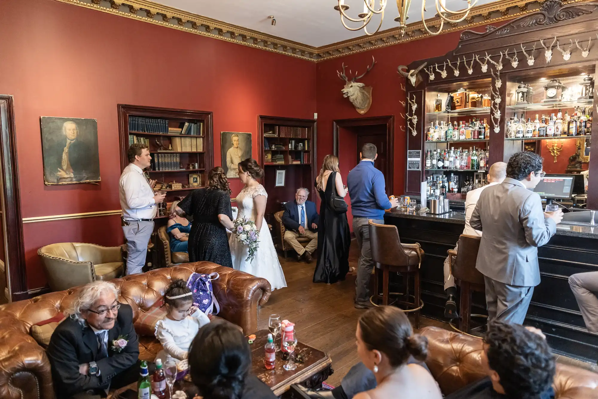 A wedding reception in a vintage-style room with red walls and leather couches. Guests socialize near a bar and bookshelves. The bride, groom, and a few guests are in conversation, while others are seated.