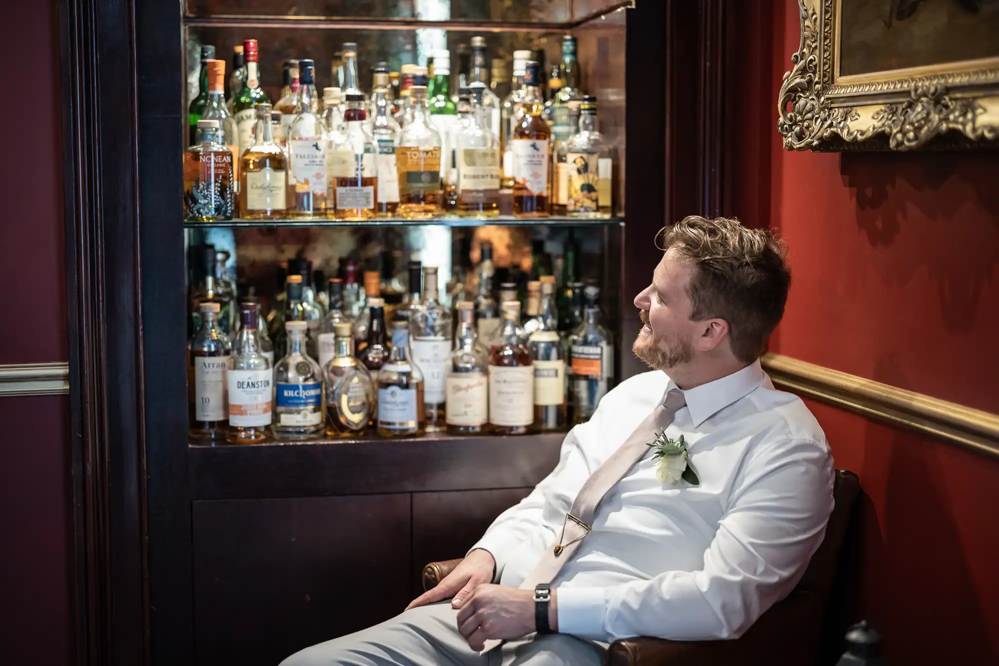 A man in formal attire sits in a chair looking at a shelf filled with various bottles of liquor.