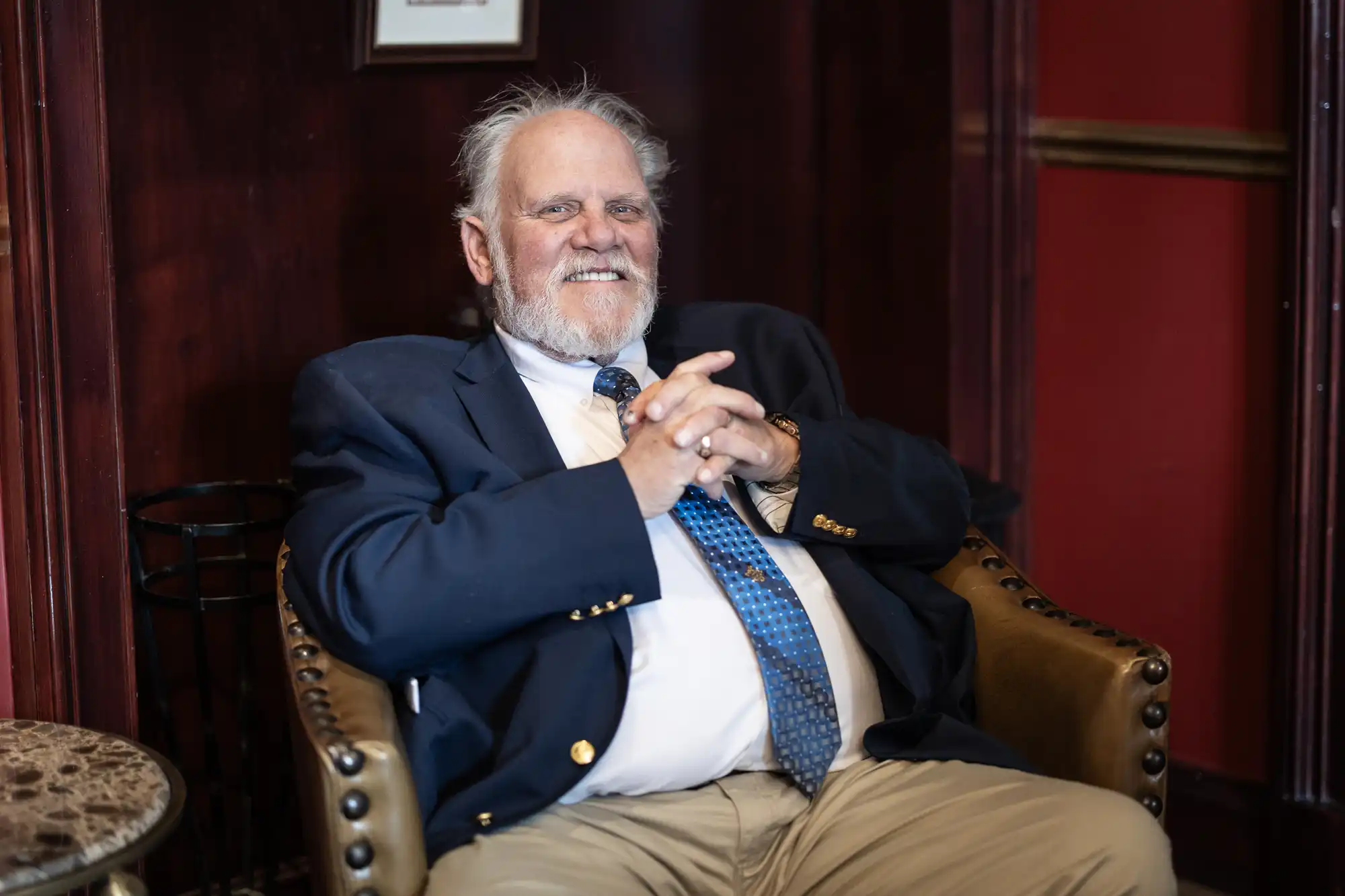 A man with white hair and a beard, wearing a navy suit jacket and blue tie, sits comfortably in a leather chair, smiling.