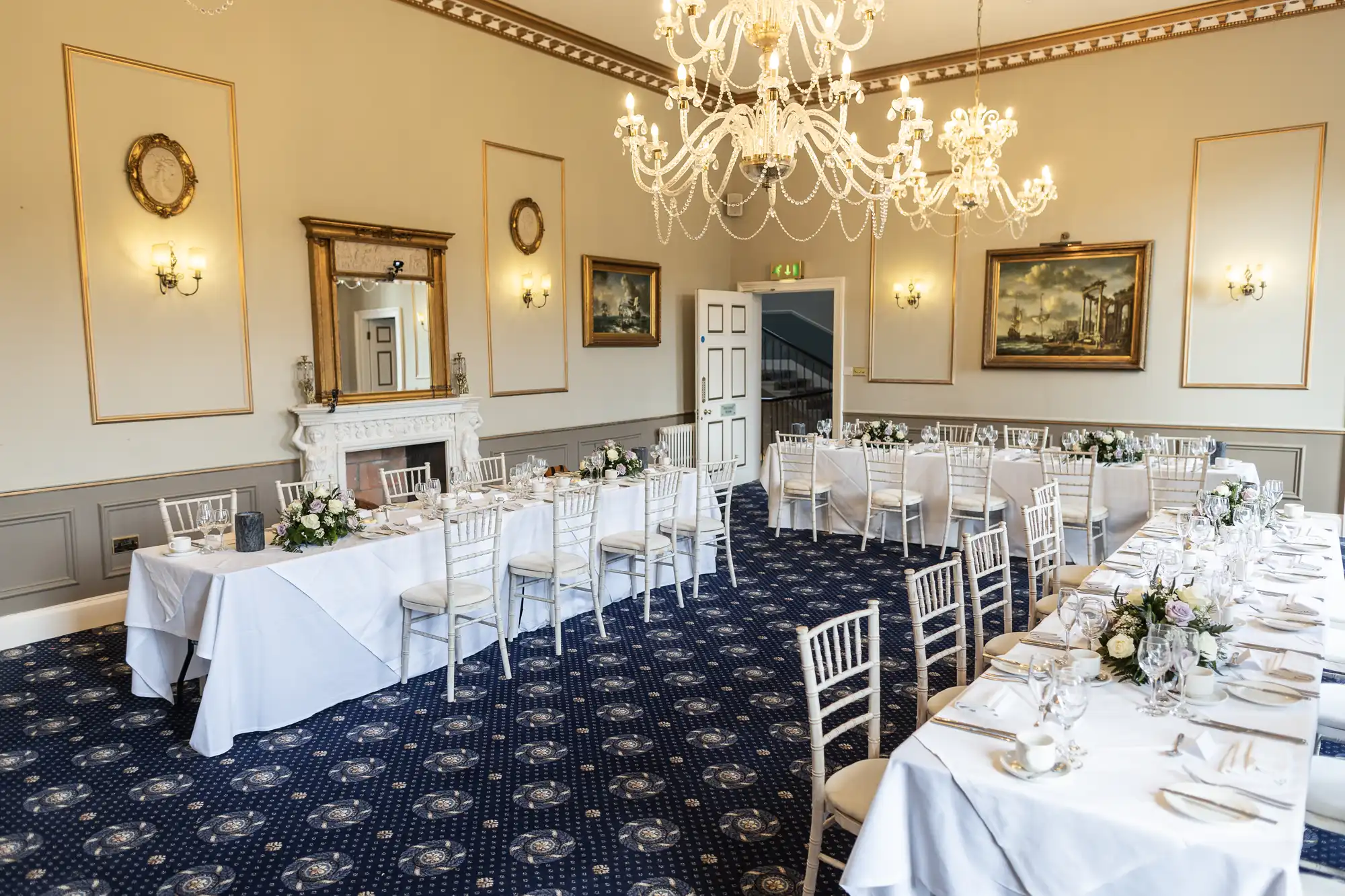 Elegantly decorated dining room with white tablecloth-covered tables, white chairs, floral arrangements, chandeliers, and framed paintings on the walls.