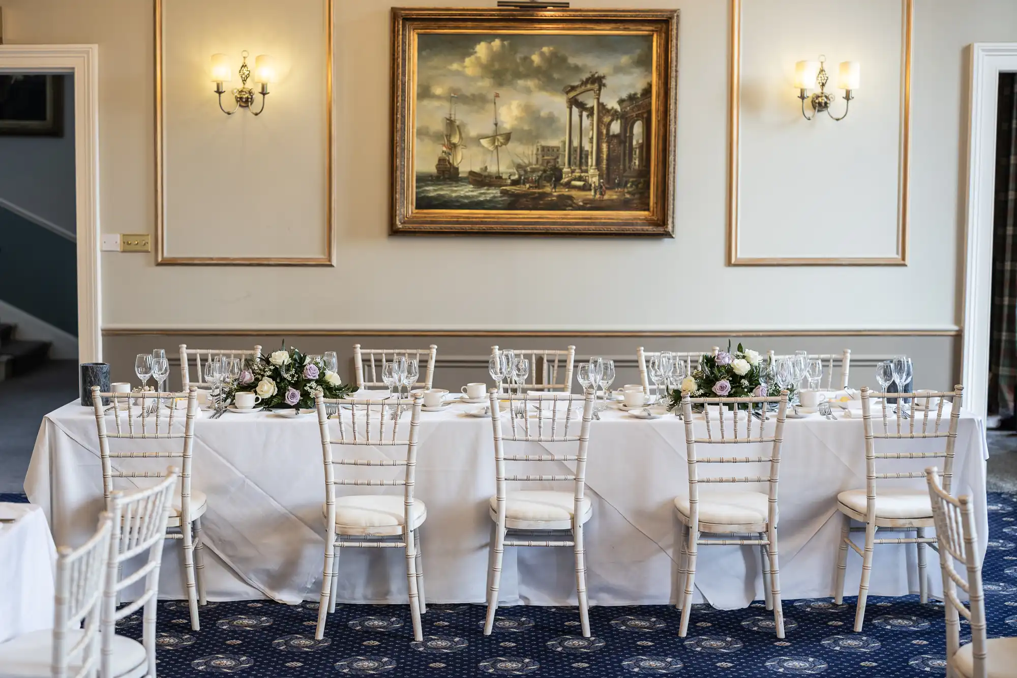 A rectangular dining table with white tablecloth, floral centerpieces, and tableware is set in an elegant room featuring a large framed painting and two wall sconces.