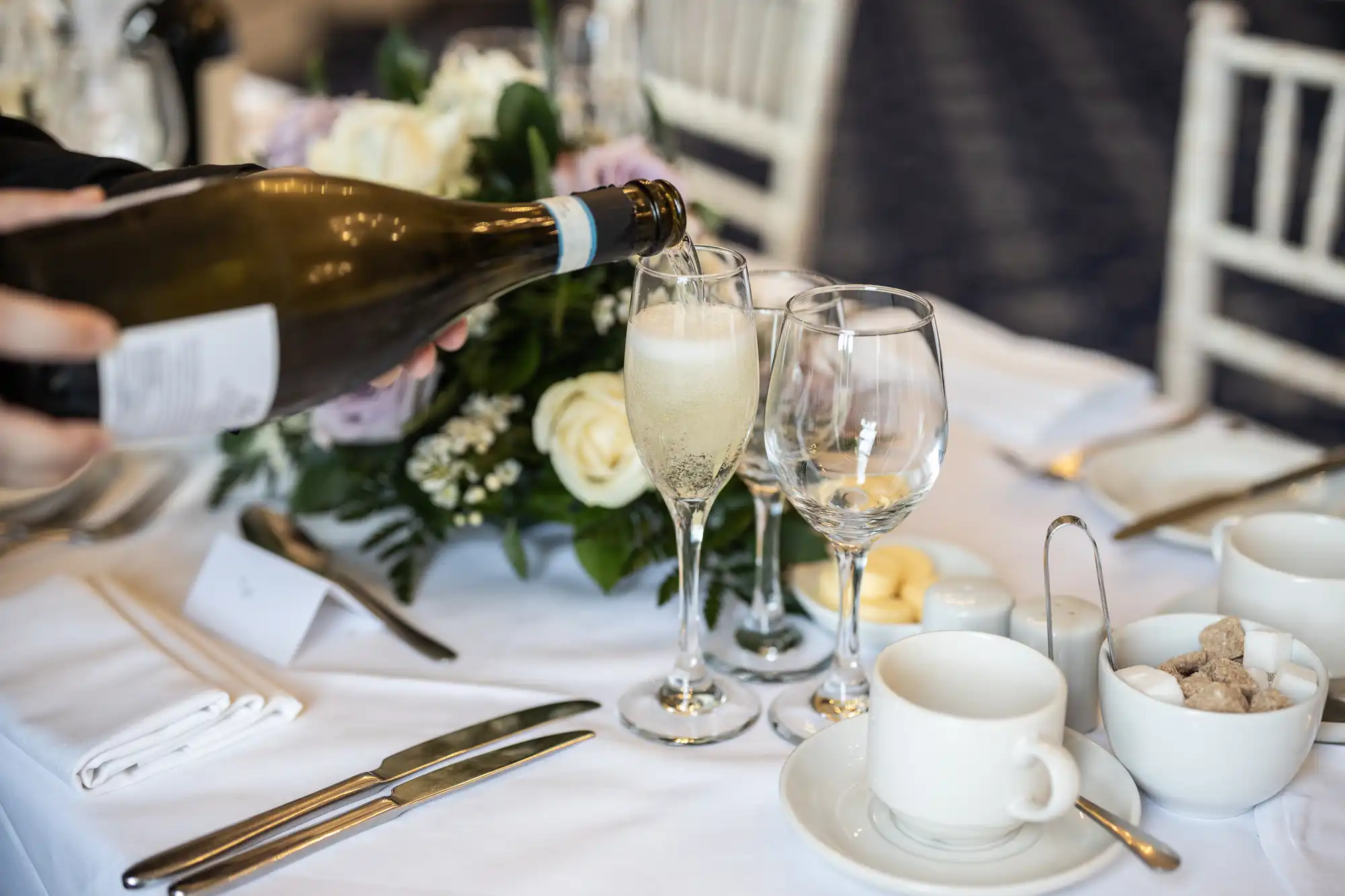 A hand pours champagne into a flute on a set dining table that includes a floral centerpiece, empty wine glasses, a teacup, cutlery, and bowls with sugar cubes and butter.