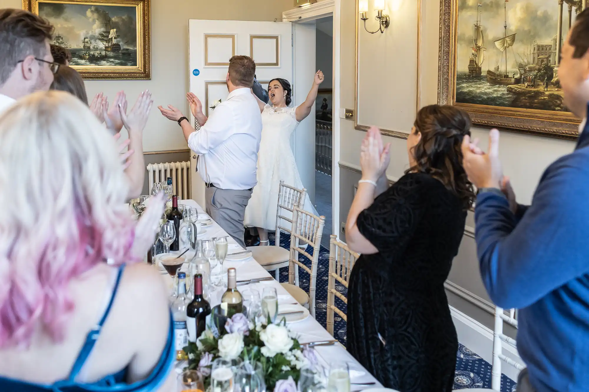 A bride and groom enter a room filled with cheering guests seated at a long dining table set for a meal, with vintage ship paintings on the walls.