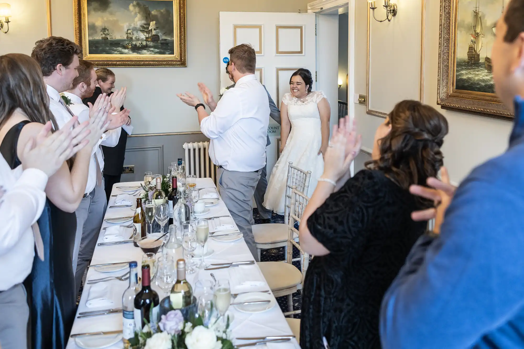 A bride enters a dining room as wedding guests clap. The room features a long table set for a meal, adorned with flowers and wine bottles, and is decorated with maritime-themed paintings.