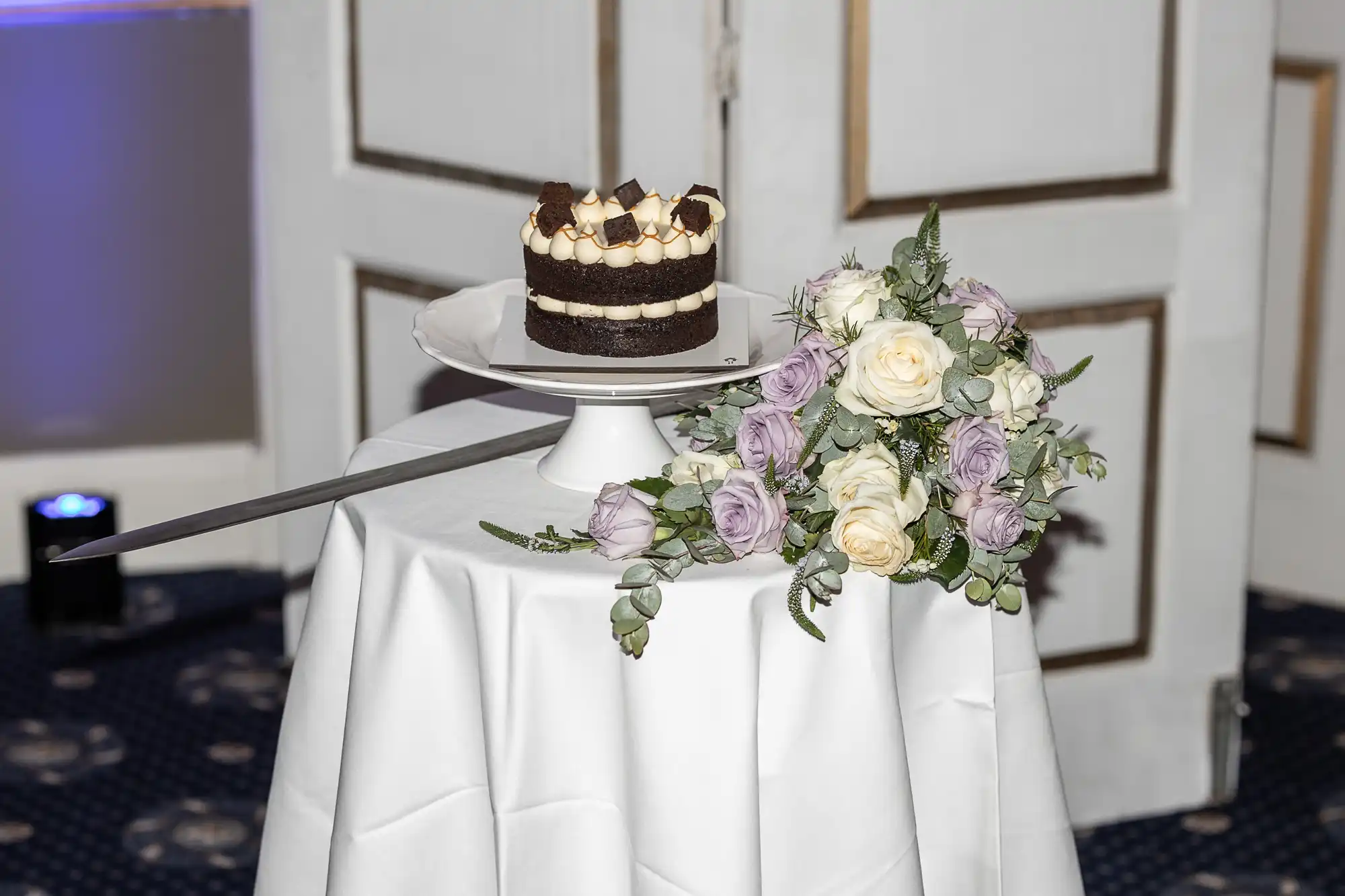 A small chocolate cake on a stand and a bouquet of white and lavender roses are displayed on a round, white-clothed table.