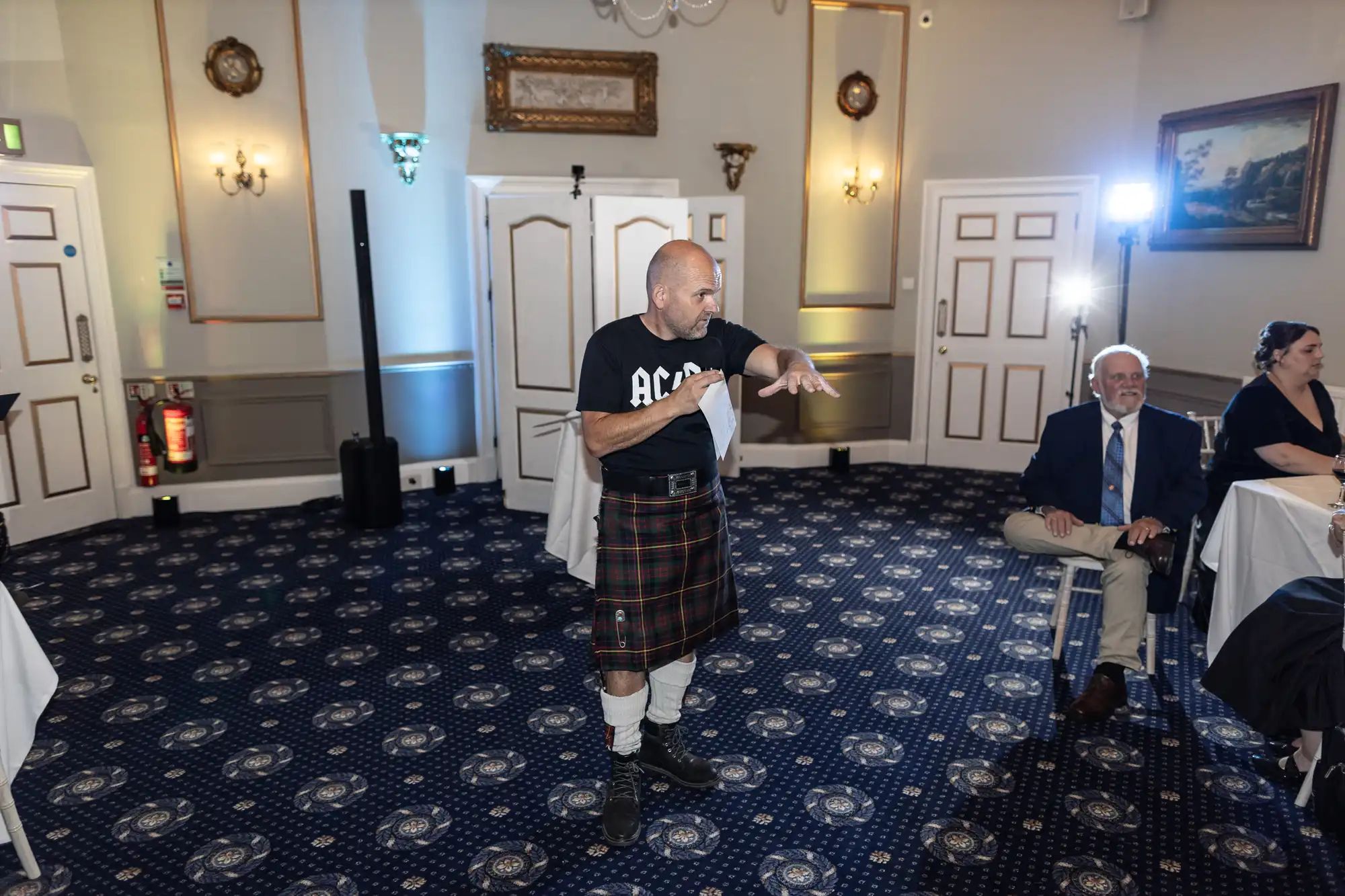 A man in a kilt and an AC/DC t-shirt points while standing on a carpeted floor in a formal room. Seated individuals watch him, and bright light shines from a corner of the room.