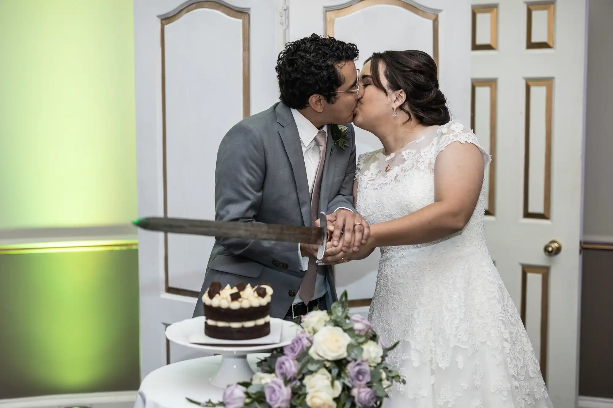 A bride and groom kiss while holding a sword together, preparing to cut a chocolate cake with flowers in the foreground.