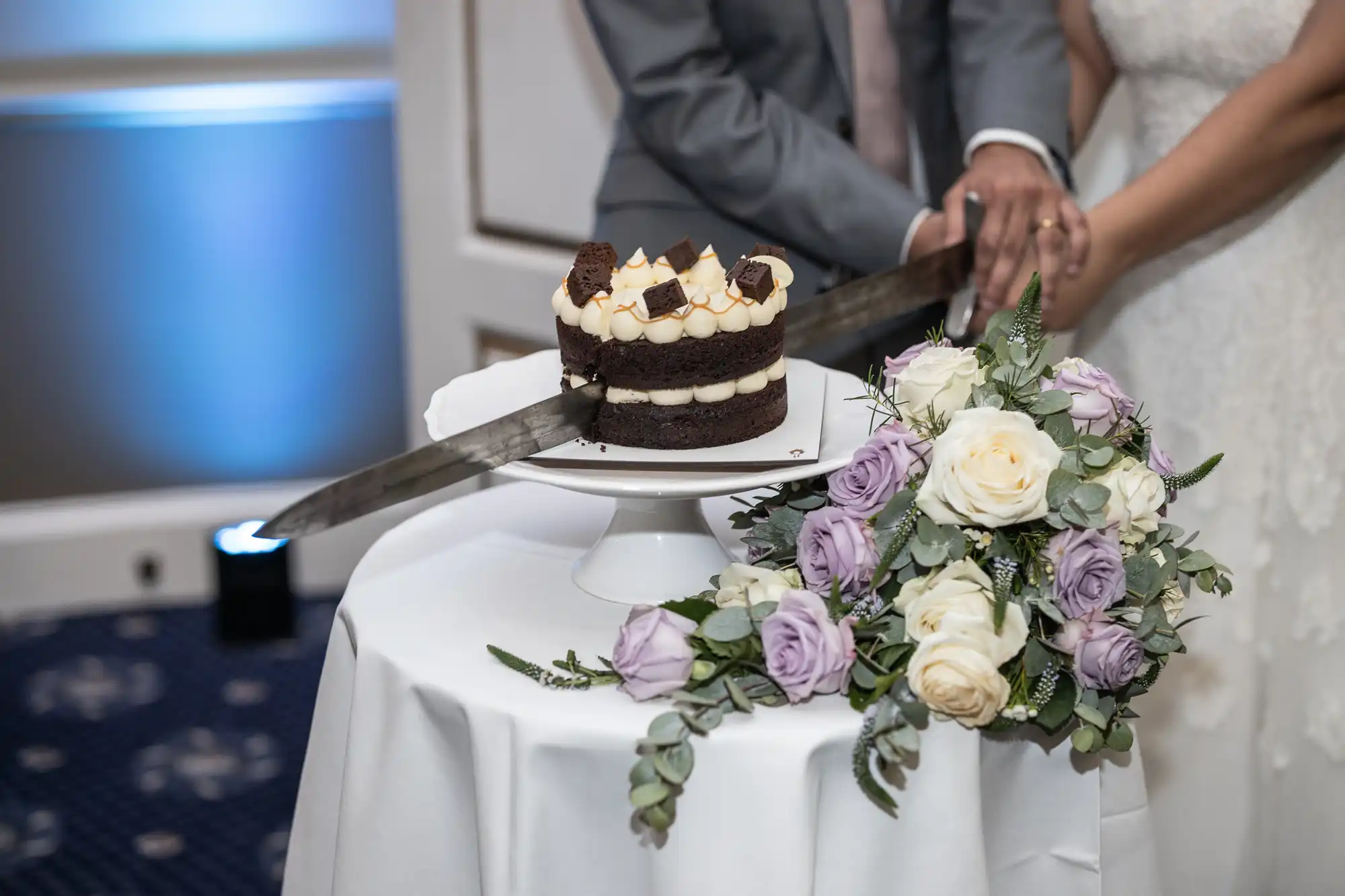 A couple cuts a small, multi-layered cake with a sword on a white table next to a bouquet of white and lavender roses.