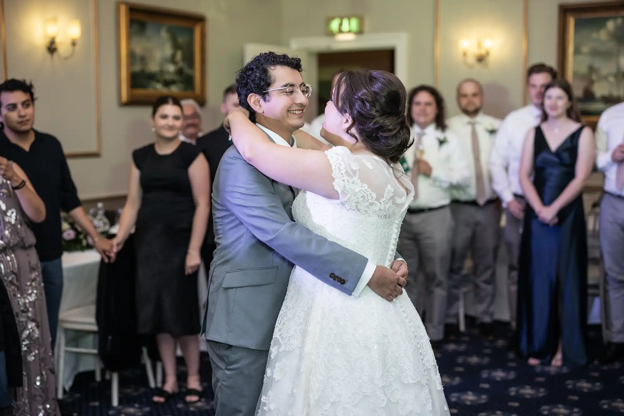 A bride and groom embrace and smile during their first dance at their wedding, surrounded by guests who are standing and watching.