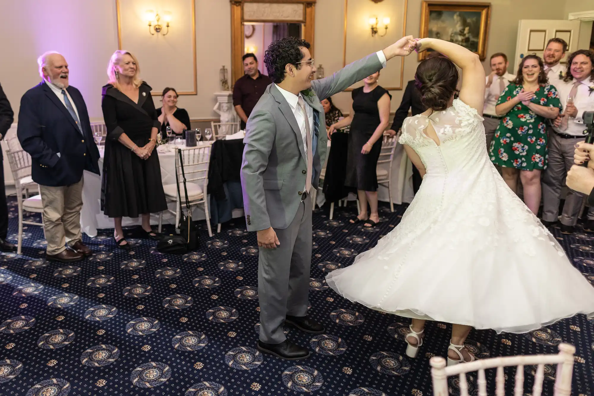 A couple dances in the center of a room as guests watch and smile. The man is wearing a gray suit, and the woman is in a white dress. Tables and chairs are set up around them.